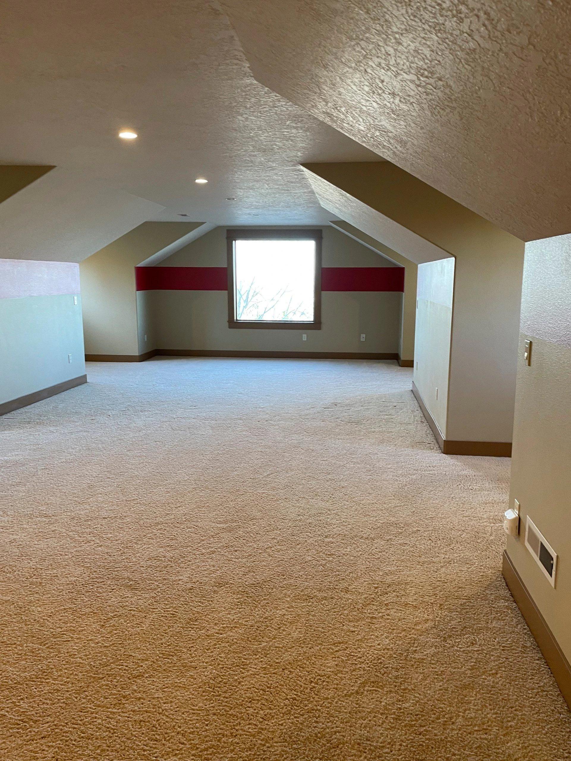 An empty carpeted room has a sloped, textured ceiling, recessed lights, and a window centered on the far wall.