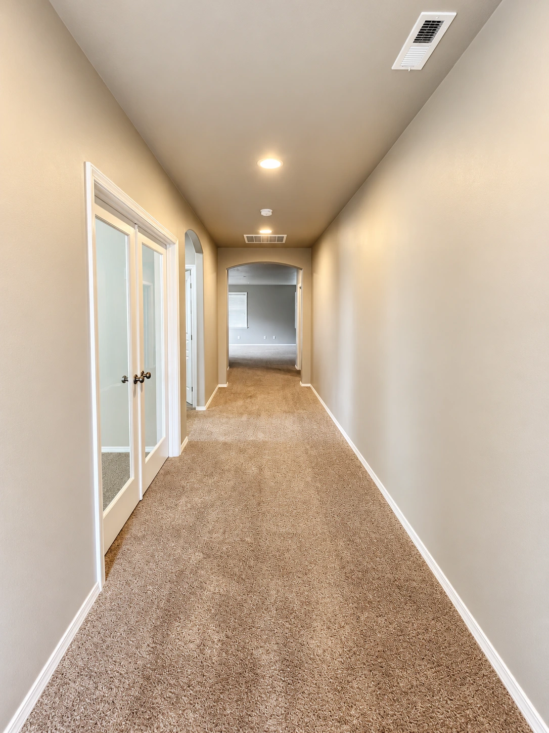 A carpeted hallway with beige walls, glass-paneled double doors on the left, and an arched opening at the far end.