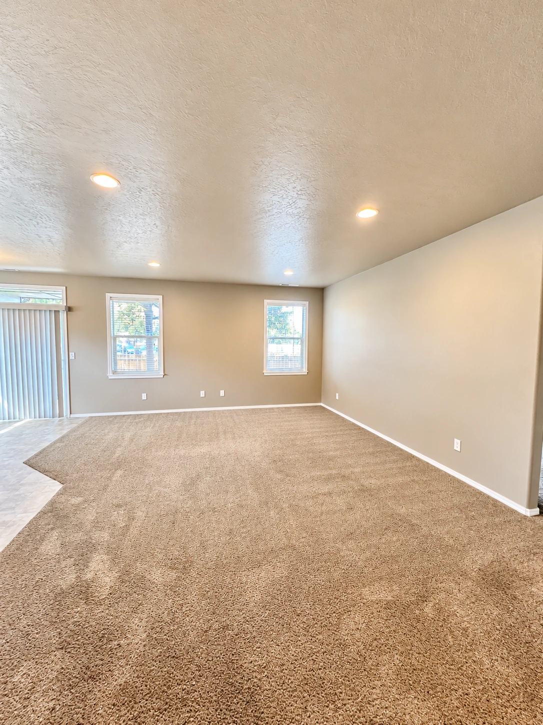 An empty room has beige carpet, tan walls, two windows, vertical blinds, and recessed ceiling lights.