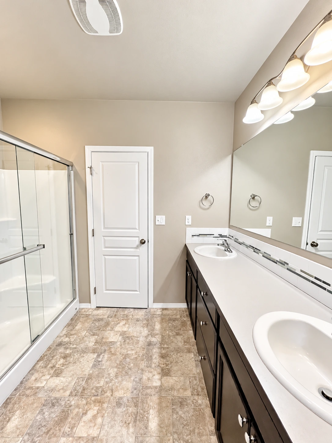 A bathroom with a glass shower, a long double-sink vanity beneath a large mirror, and patterned tile flooring.