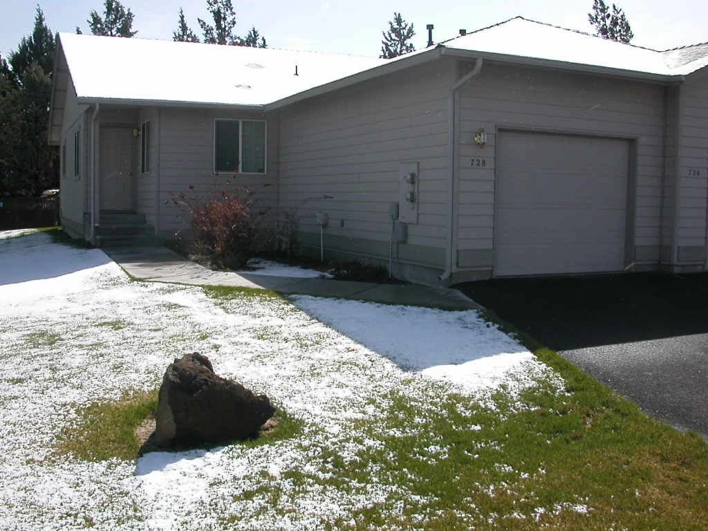 A gray house with a garage stands behind a lawn with a large rock in the foreground.