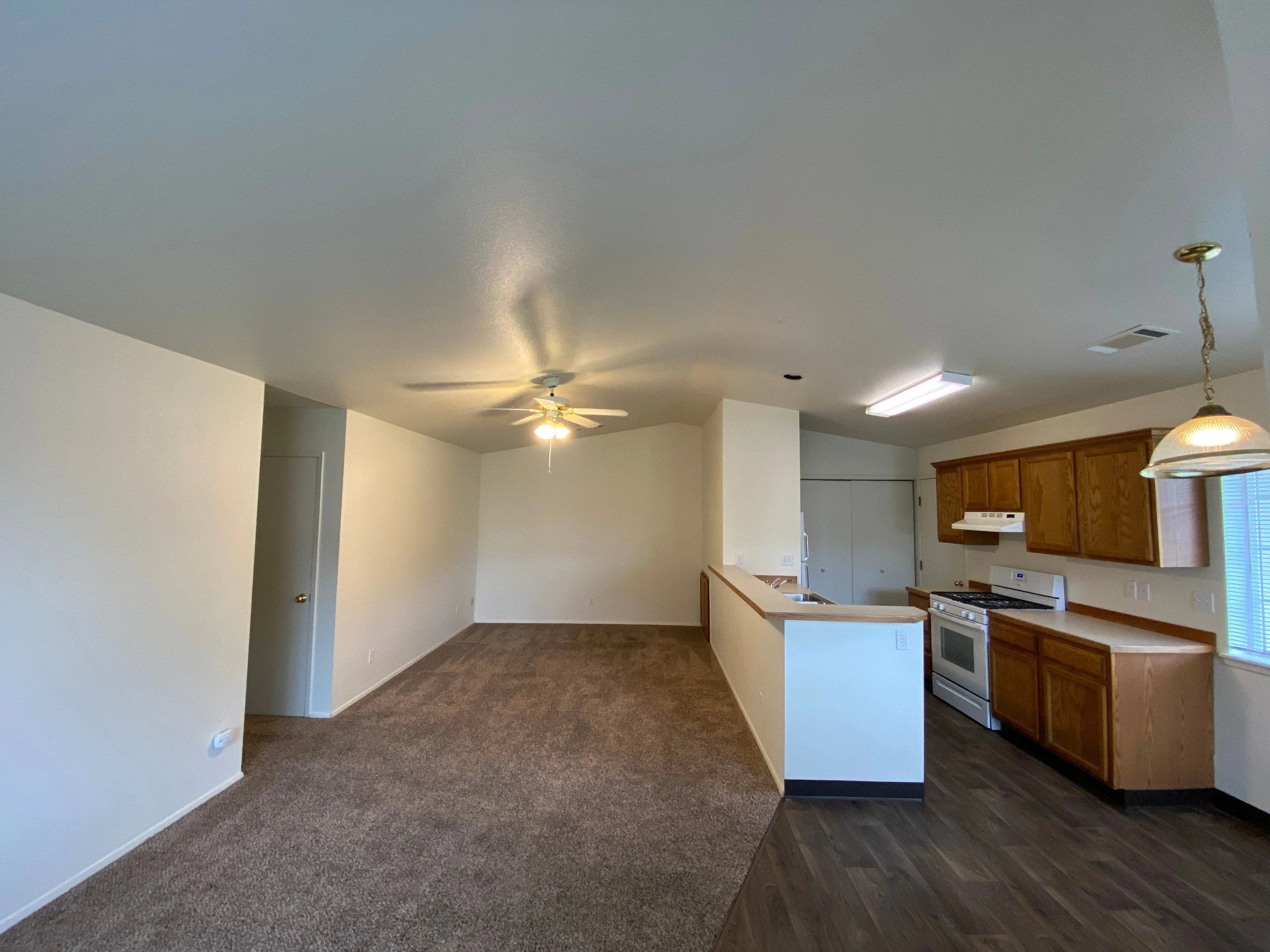 A carpeted room opens into a kitchen with wood cabinets, a counter peninsula, a stove, and a ceiling fan.