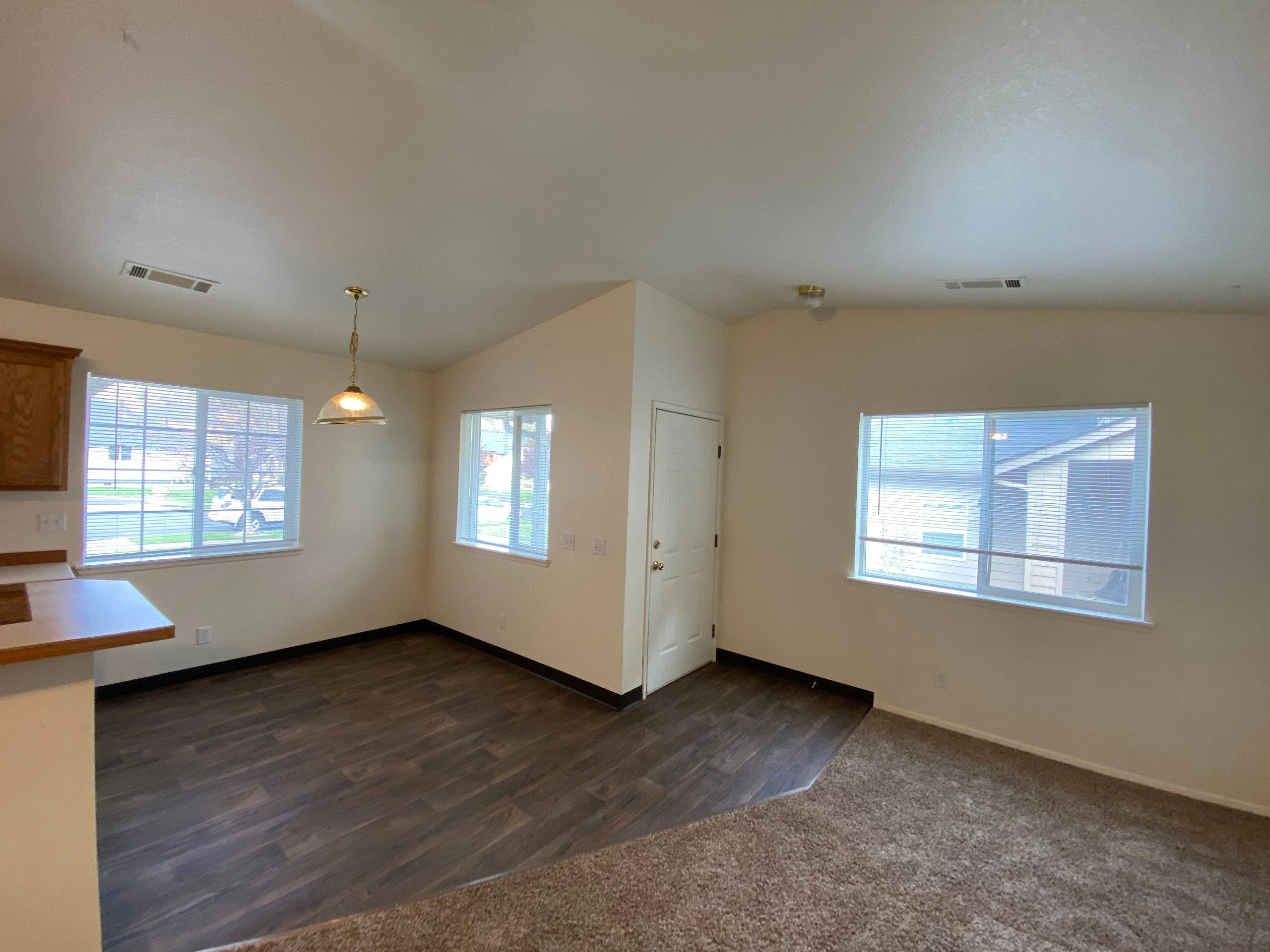 An empty room has beige walls, three windows with blinds, a white door, dark flooring, and a carpeted foreground.