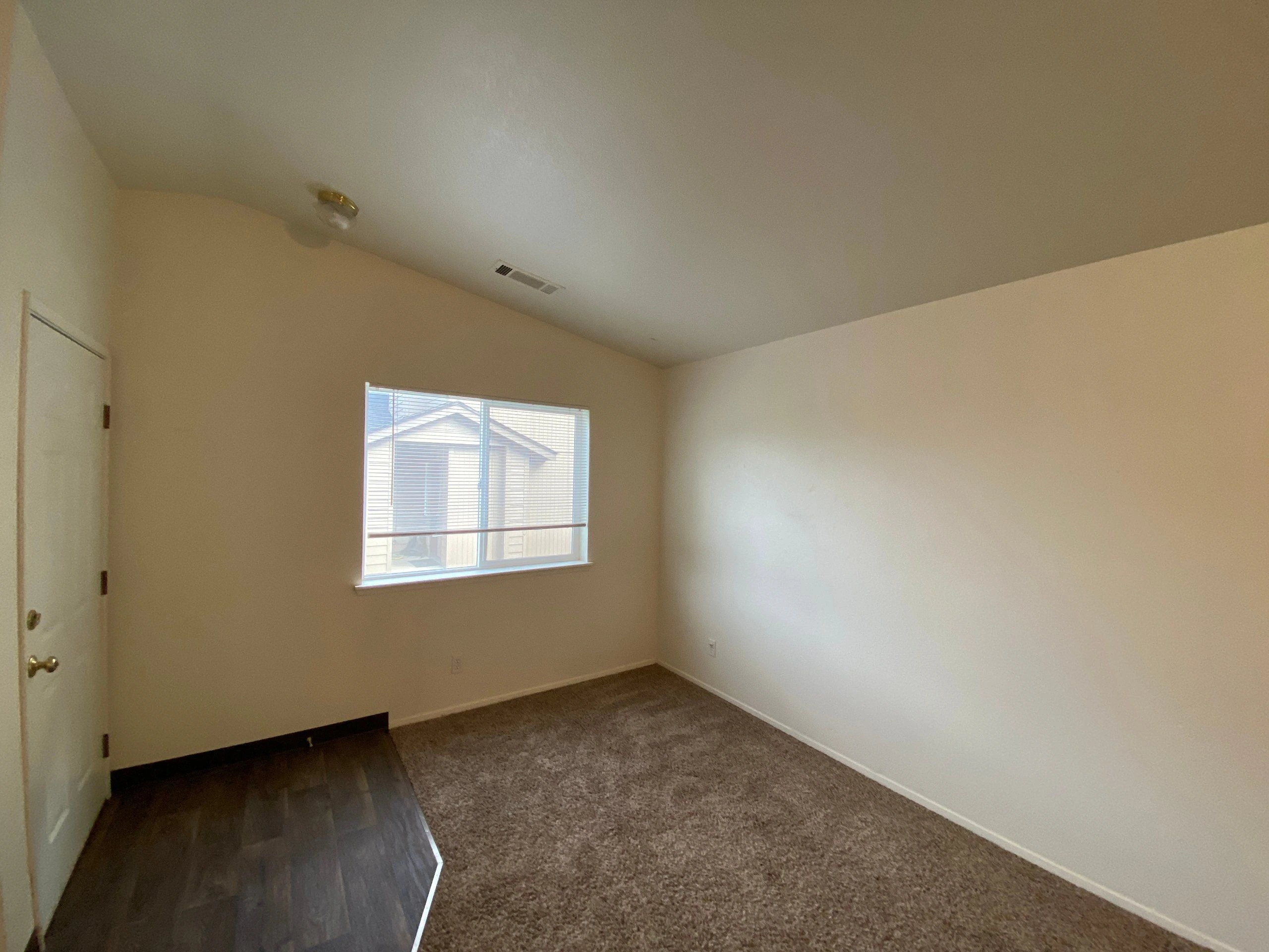 An empty room with beige walls, a window with blinds, a sloped ceiling, and carpet beside a dark hard-floor strip.