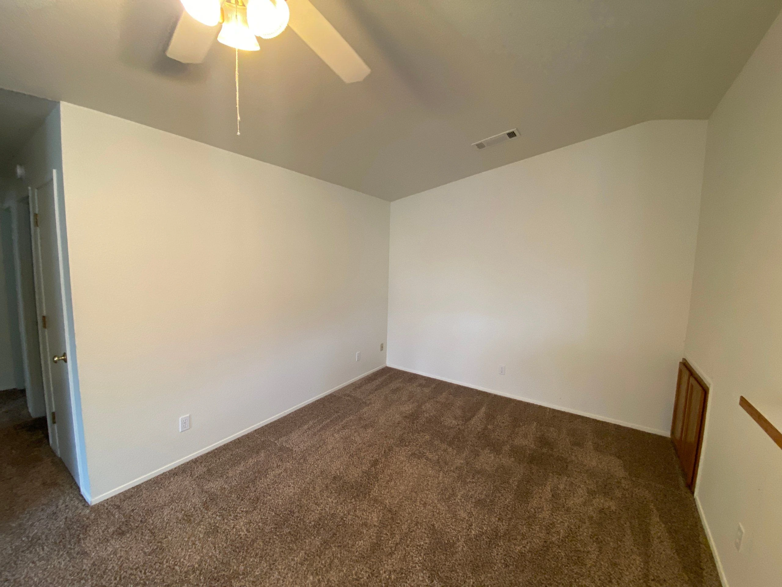 An empty room has beige walls, brown carpet, a ceiling fan with lights, and an open doorway at left.