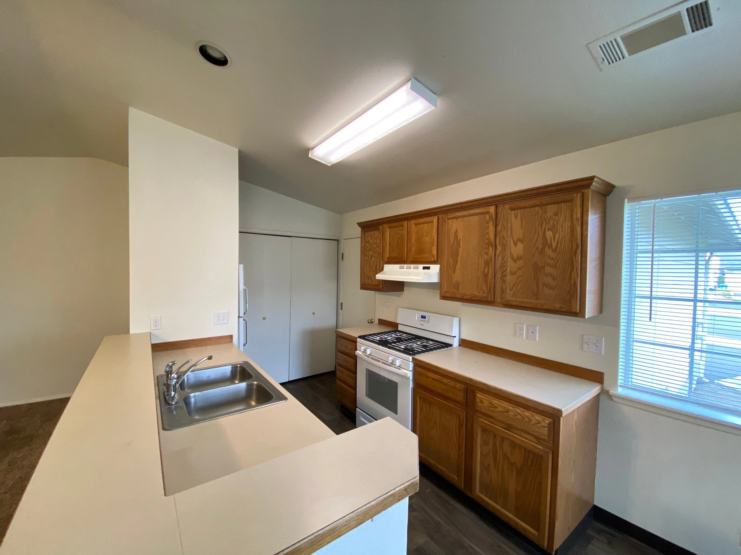 A kitchen with wooden cabinets, pale counters, a double sink, a stove, a refrigerator, and a window with blinds.