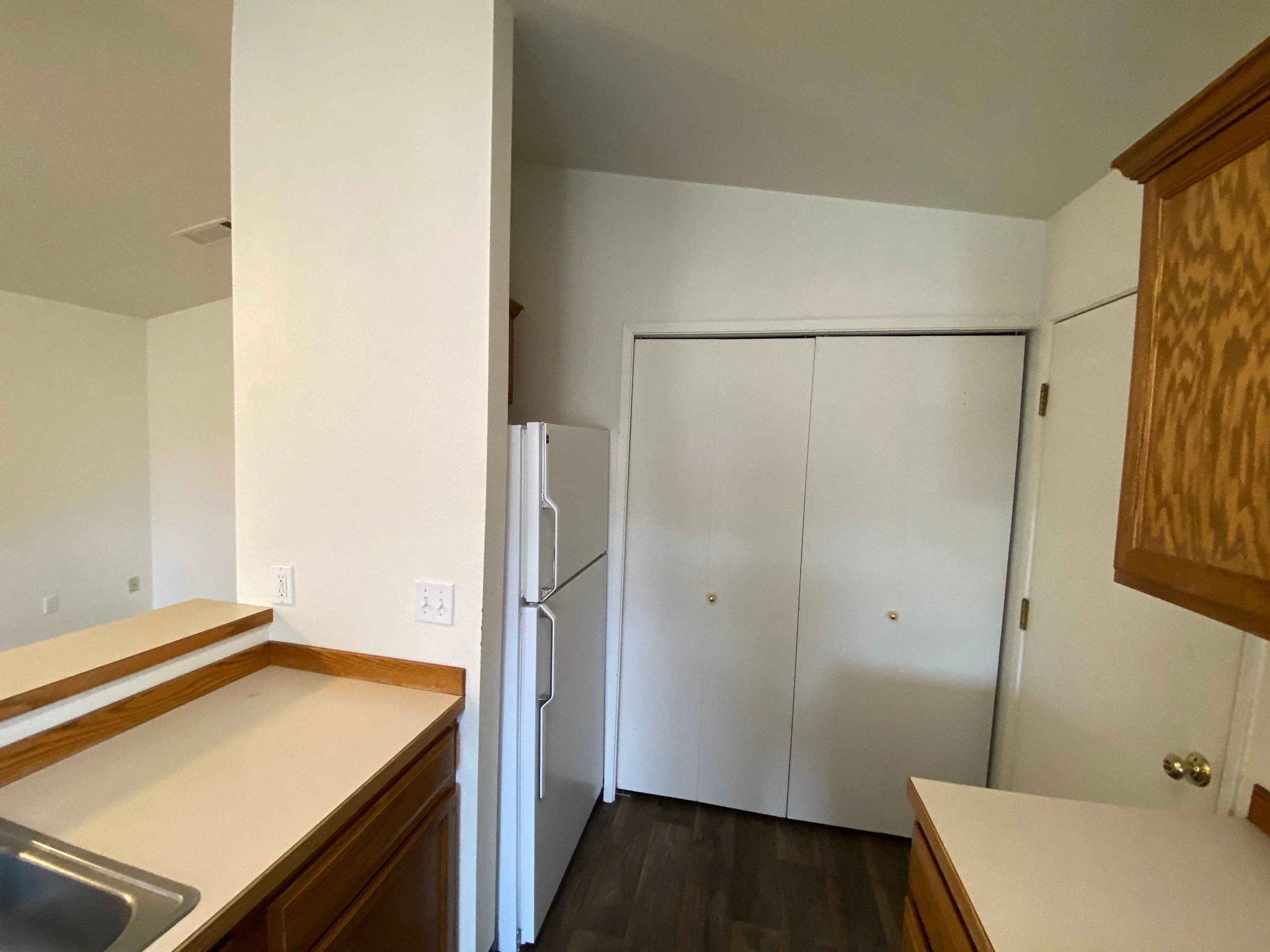 A narrow kitchen with wood cabinets, light countertops, a refrigerator, and white sliding closet doors.