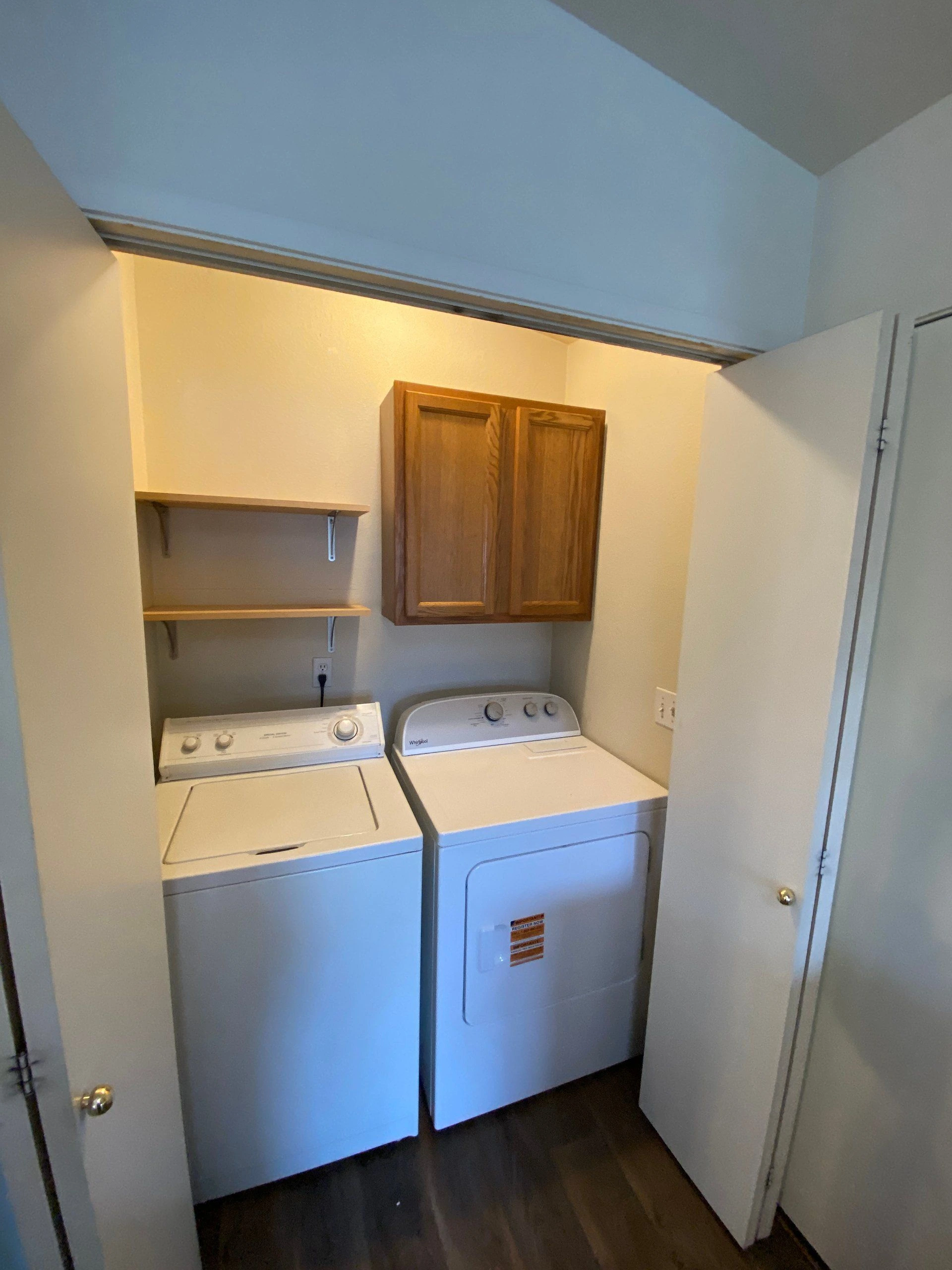 A top-loading washer and front-loading dryer sit beneath a wooden cabinet and two open shelves.