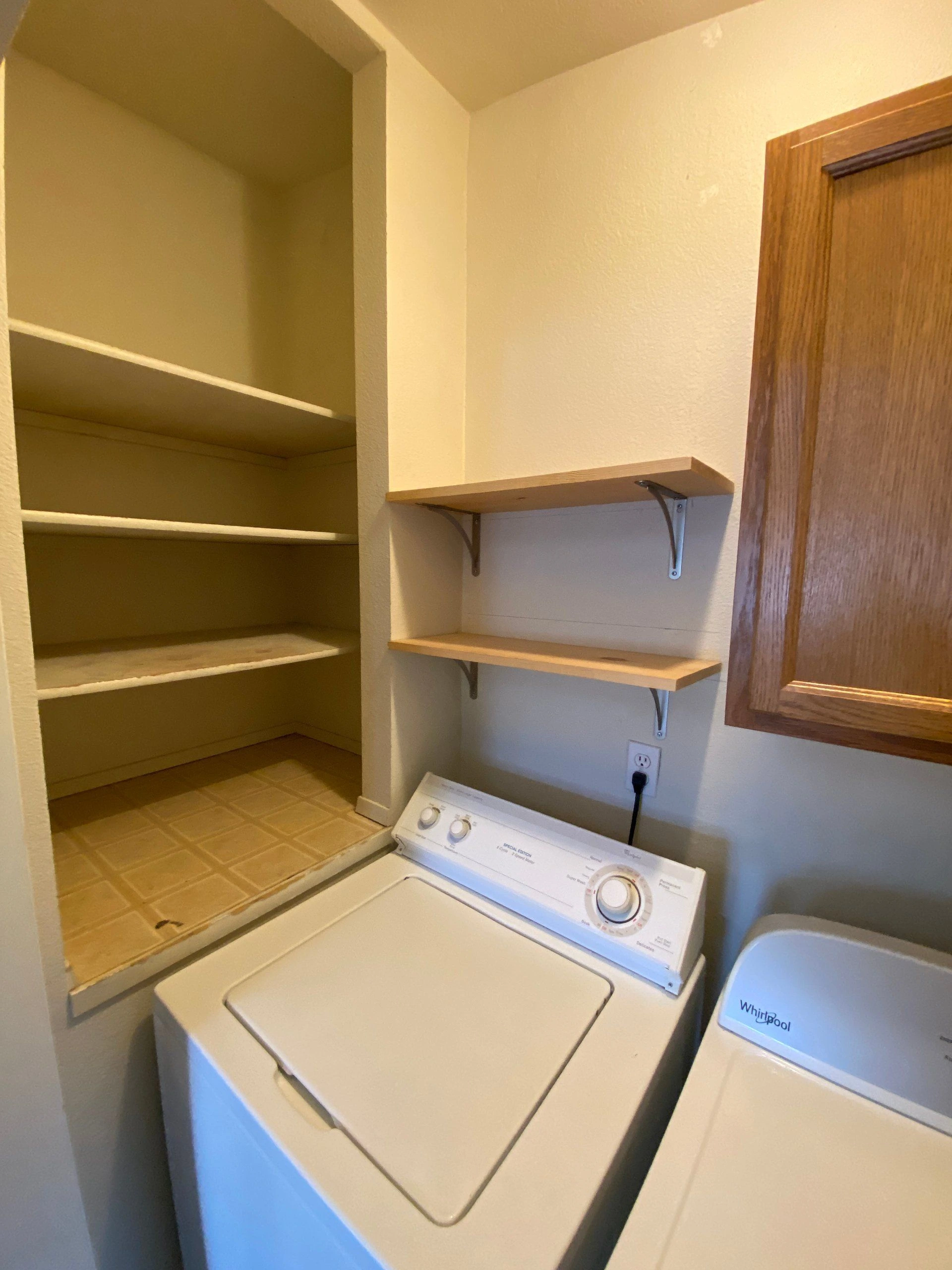 A top-loading washer and dryer sit beneath two wall shelves, beside an open shelving unit and a wooden door.