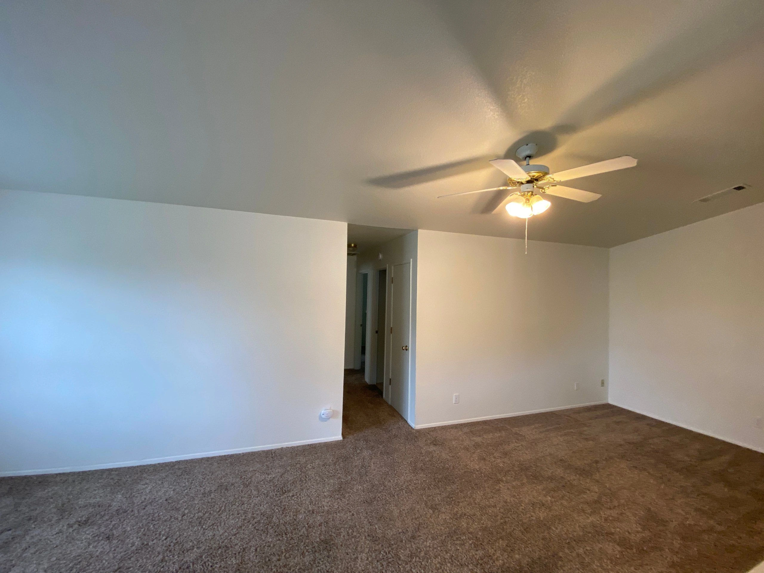 An empty room with brown carpet, white walls, and a lit ceiling fan; an open doorway leads to a dim hallway.