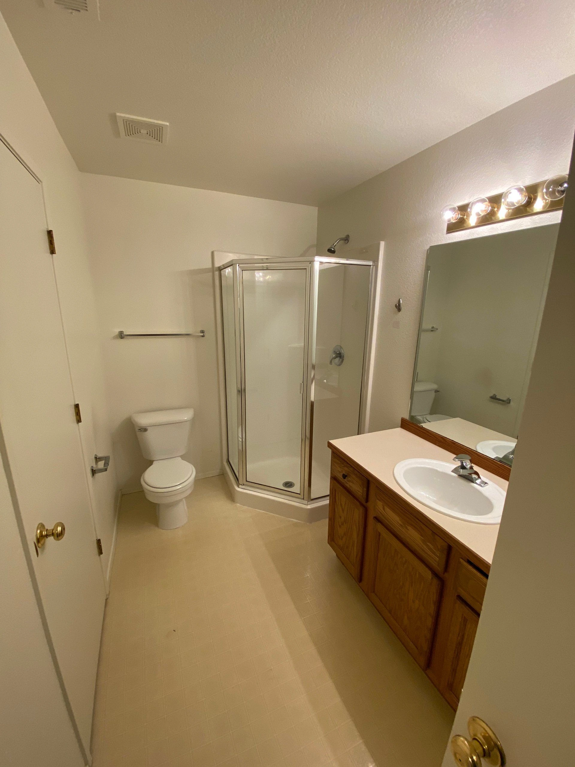 A bathroom with a glass shower enclosure, toilet, and wooden vanity with a sink beneath a large mirror and lights.