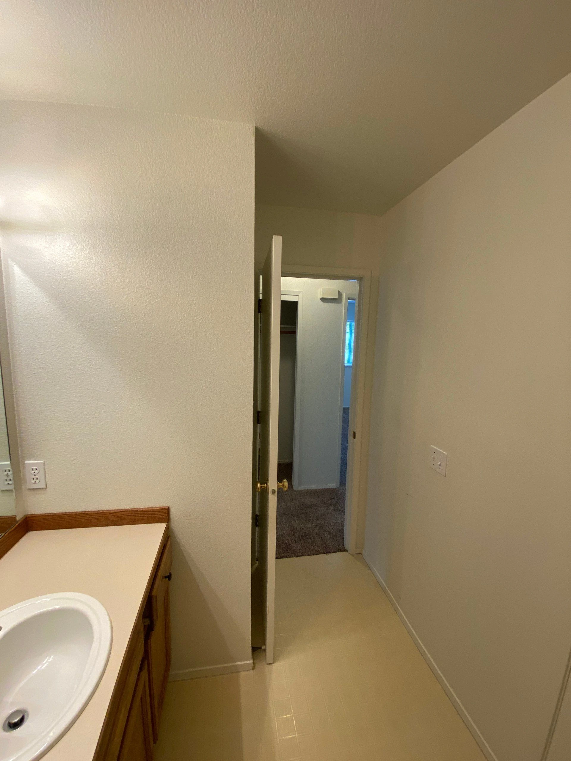A narrow bathroom has a white sink set in a wood vanity, with an open door leading to a carpeted area.