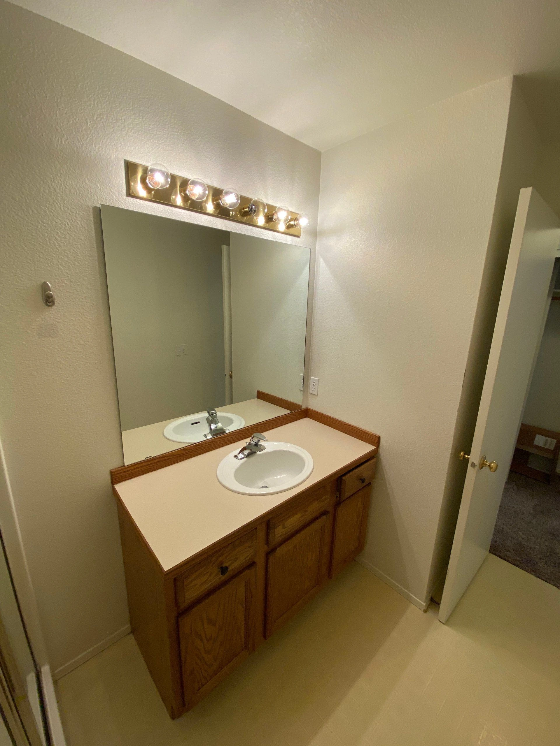 A bathroom vanity with a white sink, wooden cabinets, a large mirror, and a row of lights above it.
