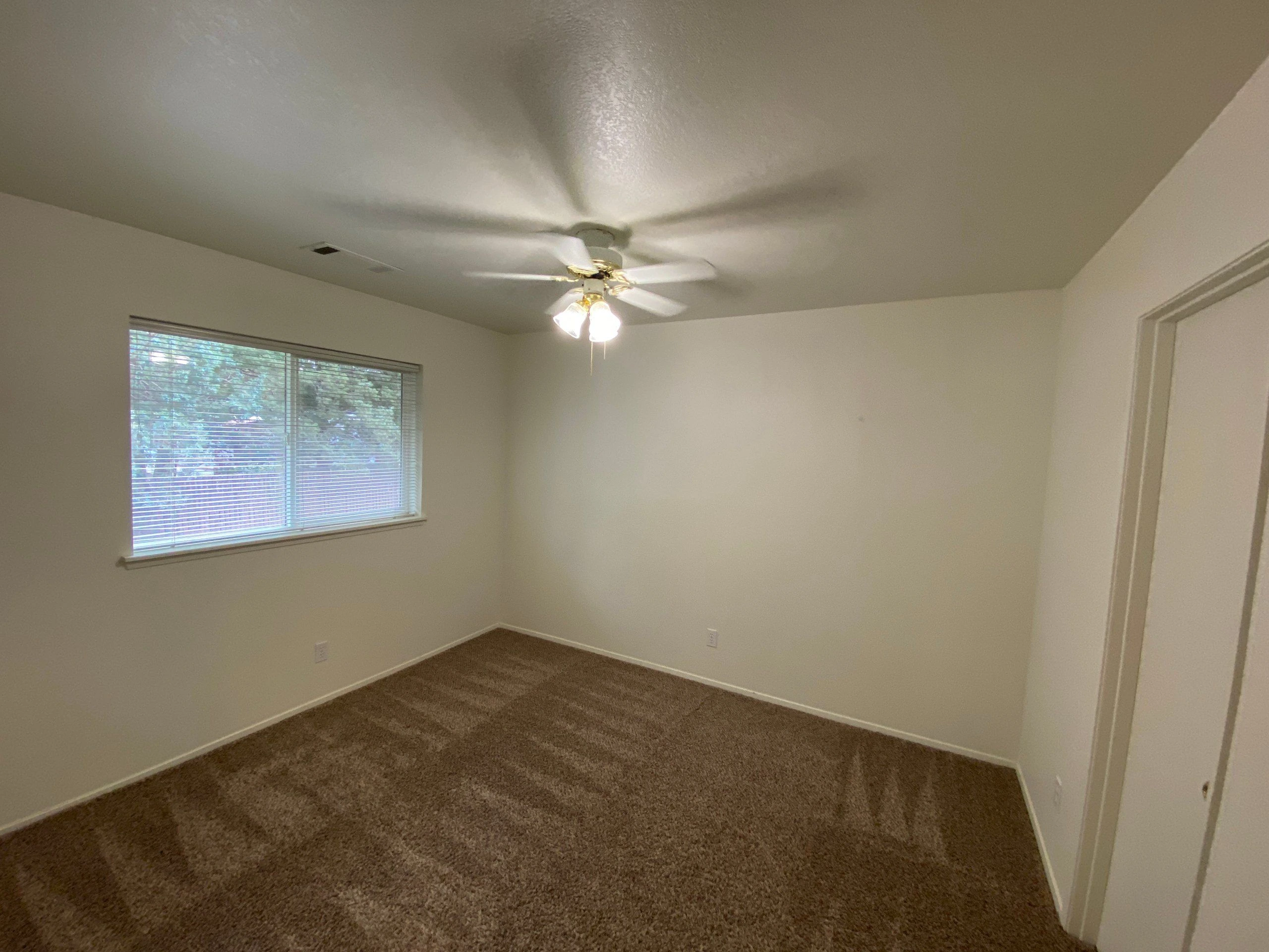 An empty room with brown carpet, pale walls, a window with blinds, and a ceiling fan with lights.