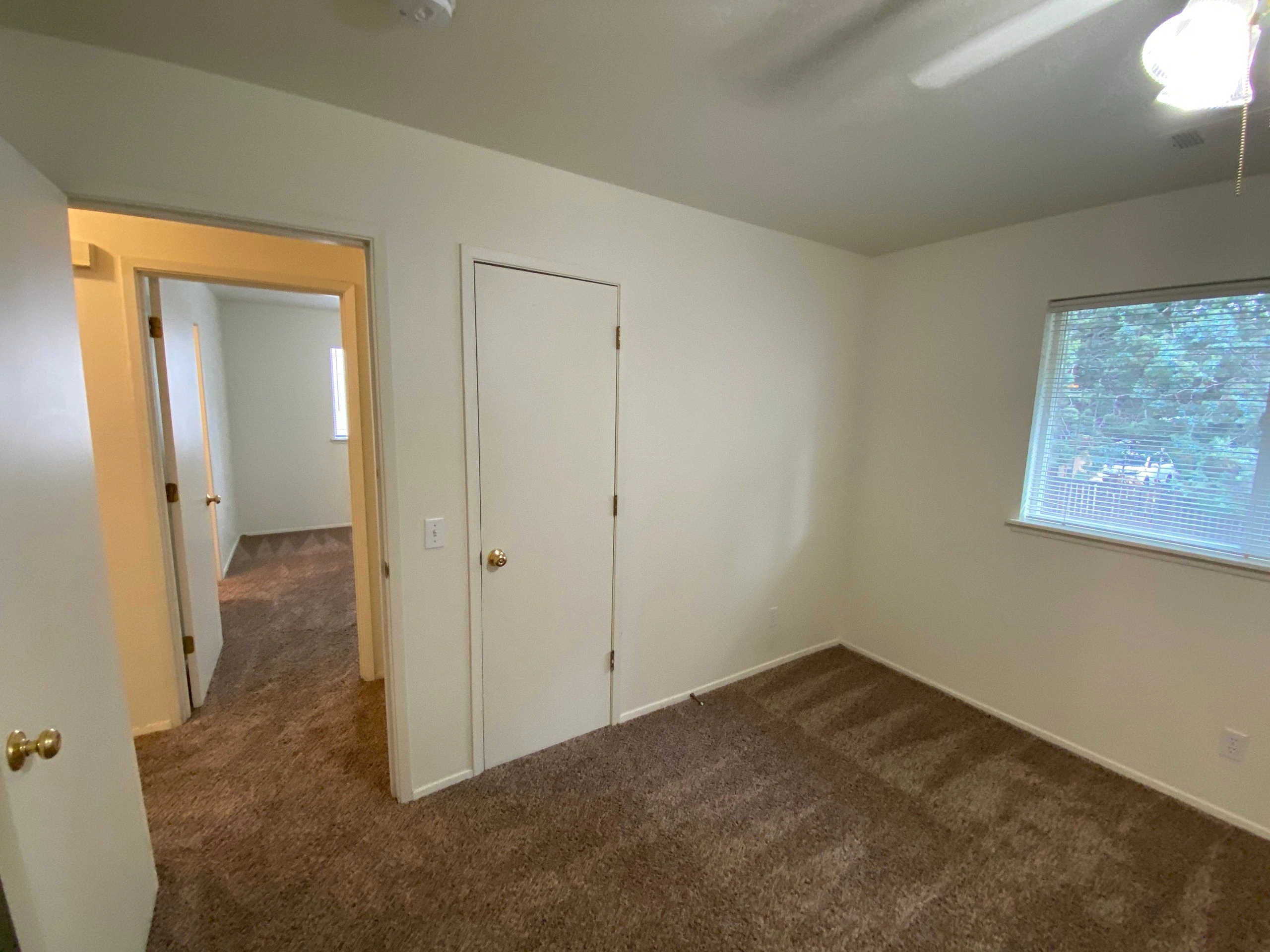 An empty carpeted room has white walls, a window with blinds, a ceiling fan, and two doors.