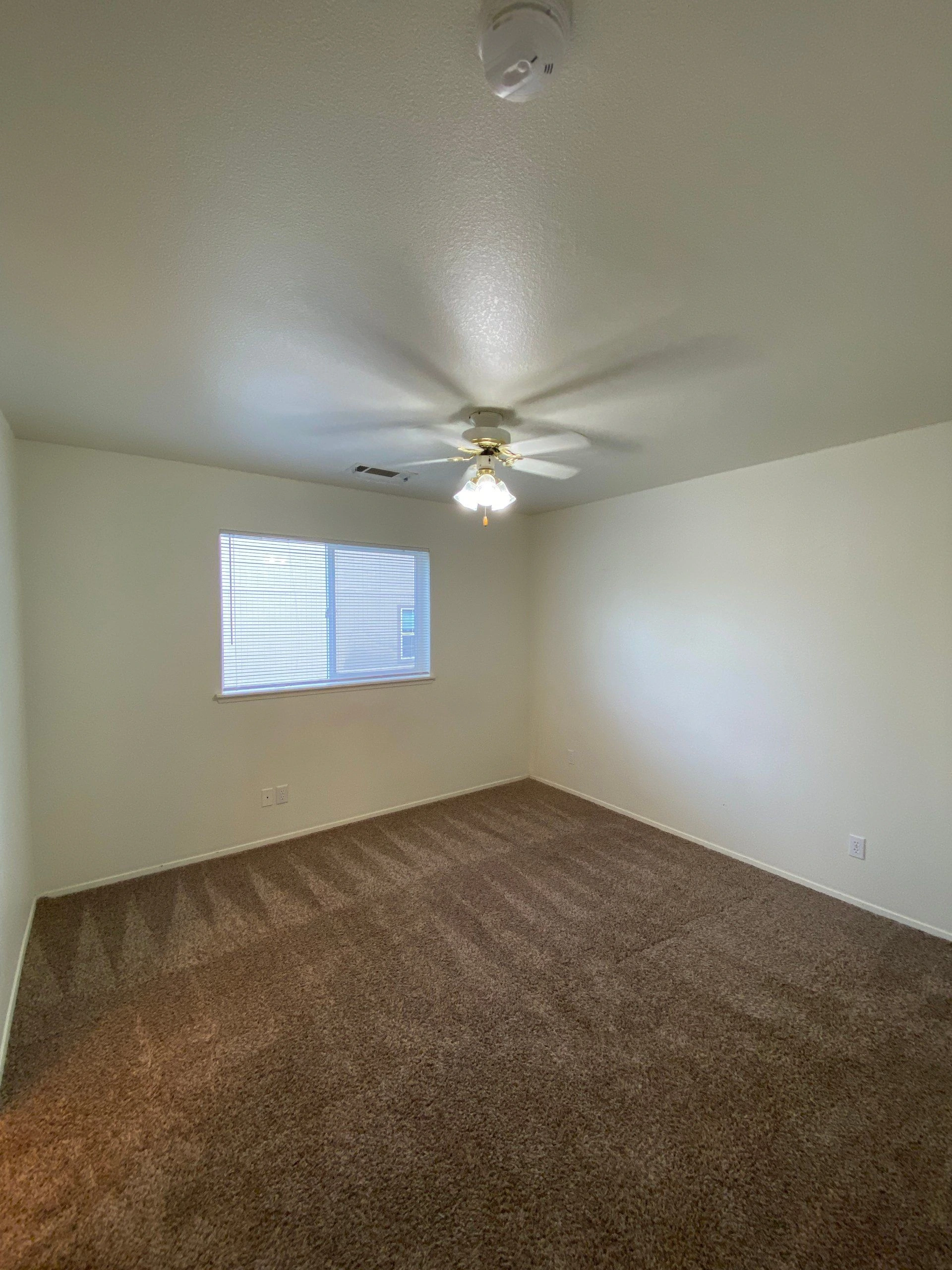 An empty room with brown carpet, pale walls, a window with blinds, and a ceiling fan with a light.