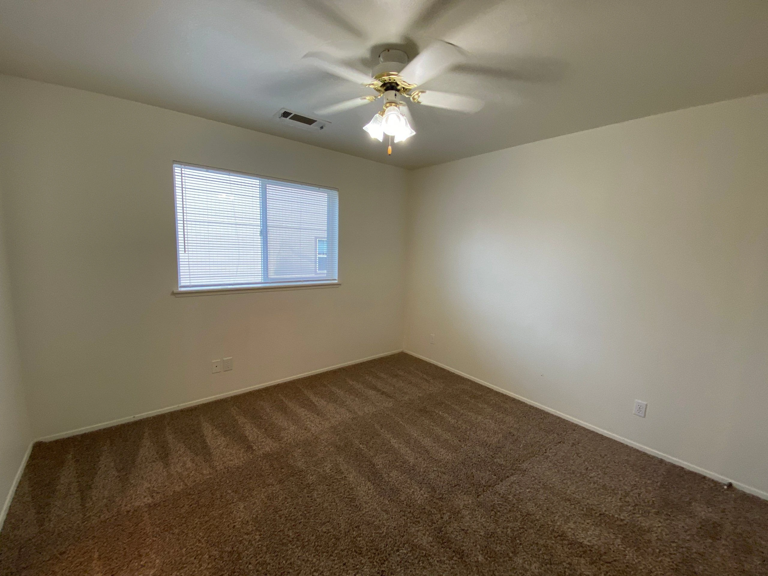 An empty room has beige carpet, light walls, a window with blinds, and a ceiling fan with a lit fixture.