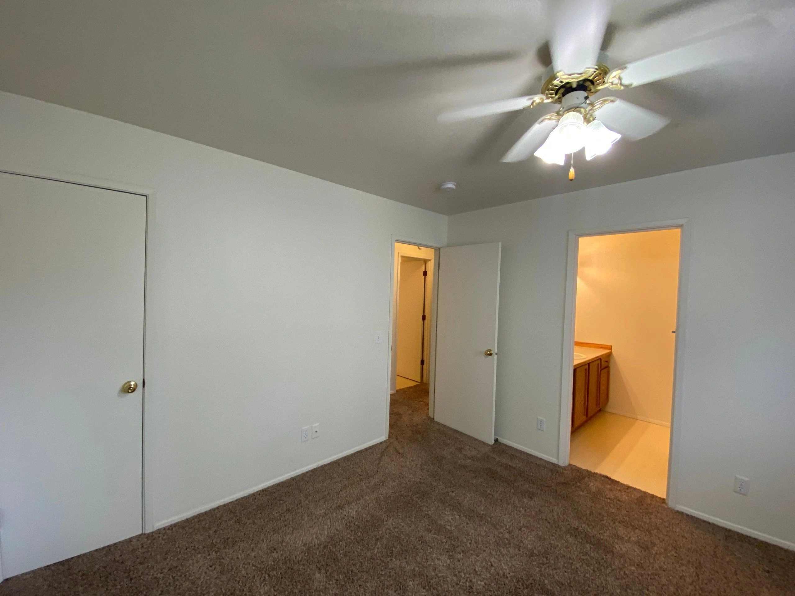 An empty carpeted room has white walls, a ceiling fan with lights, a closed closet door, and two open doorways.