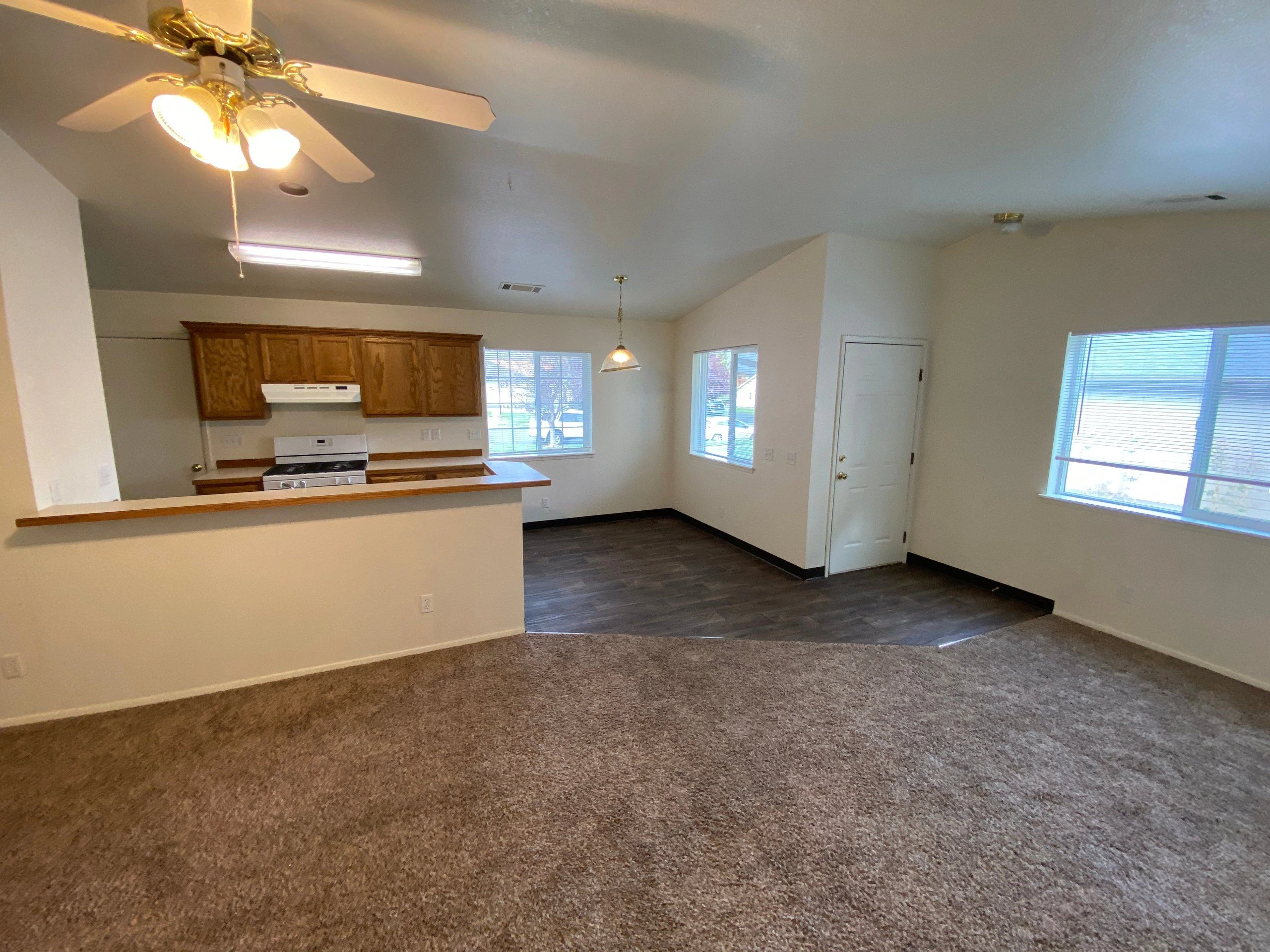 An open room has carpet in the foreground, a kitchen with wood cabinets and a counter, dark flooring, windows, and a ceiling fan.