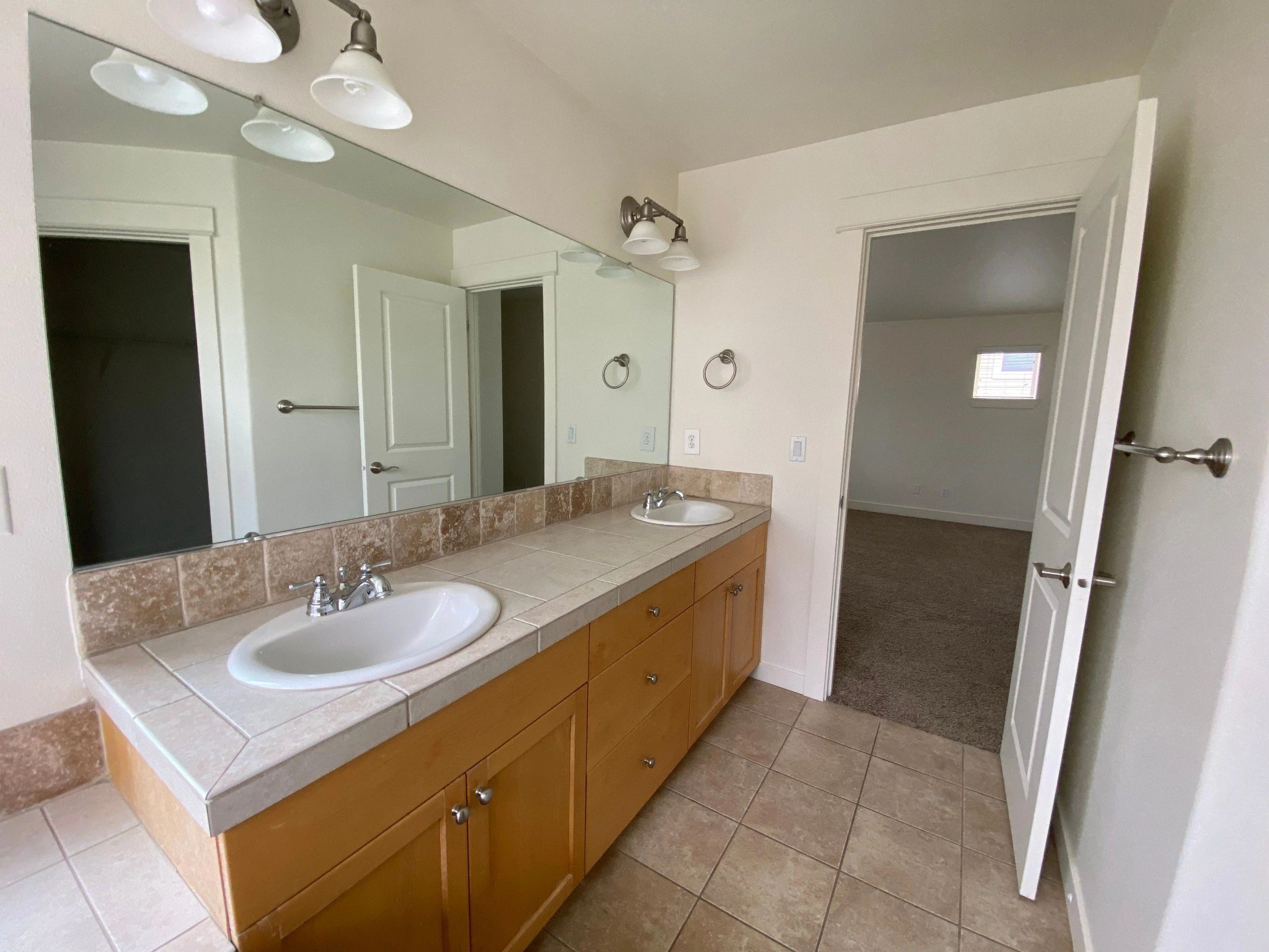 A tiled bathroom vanity has two sinks, wooden cabinets, and a large mirror.