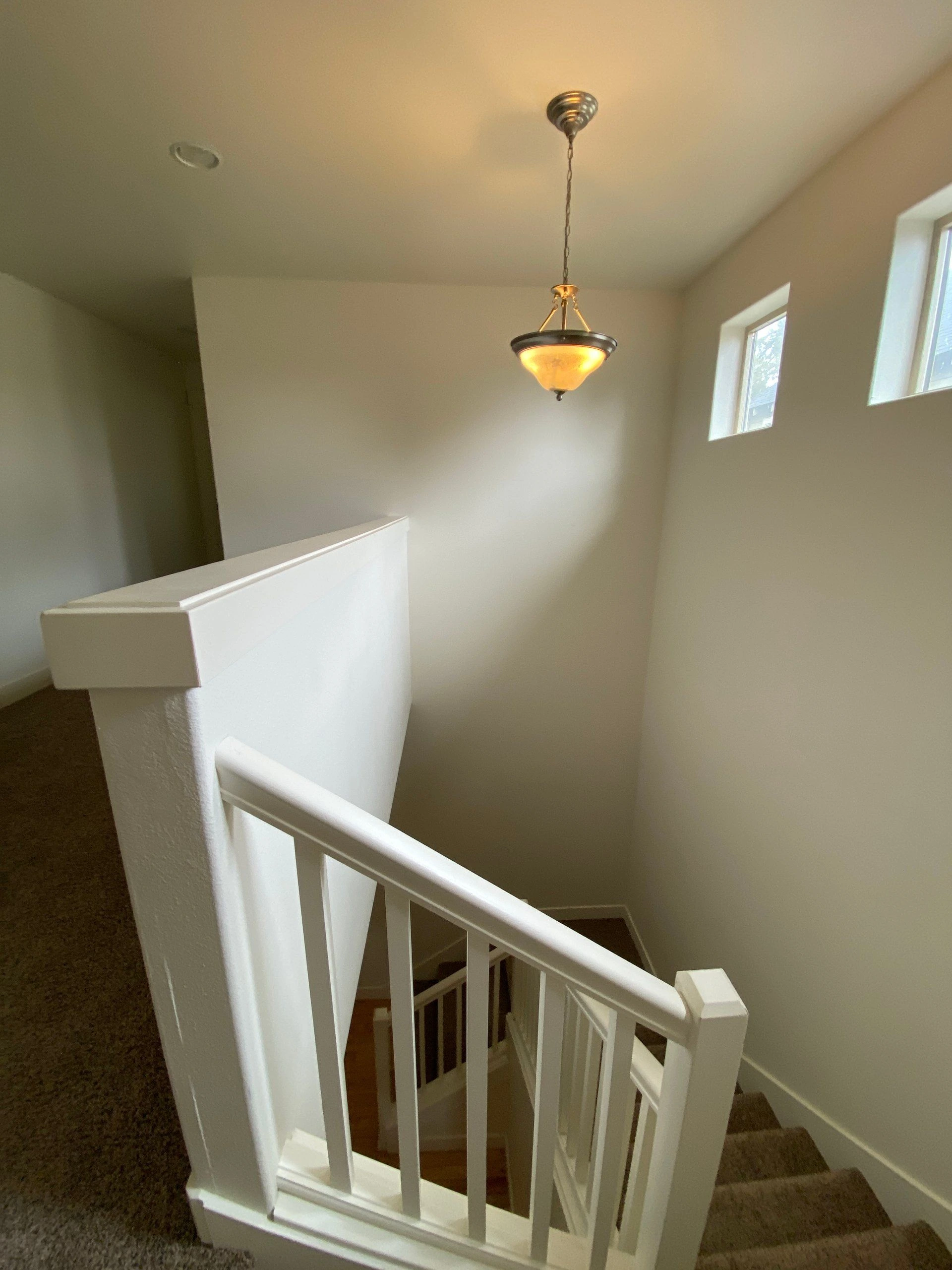 A carpeted staircase descends beside white railings beneath a hanging light, with two small windows on the wall.