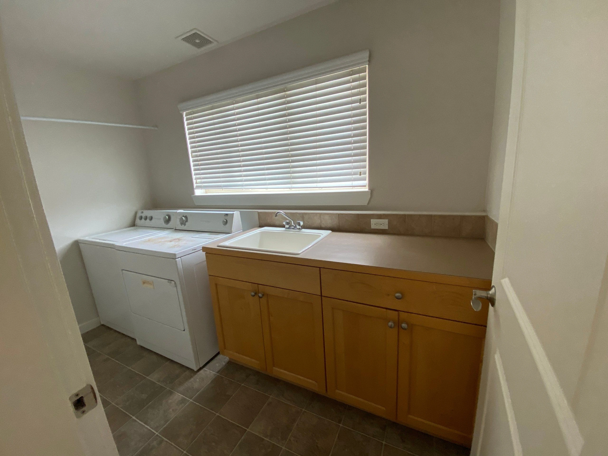 A washer and dryer stand beside a sink and wooden cabinets beneath a window with closed blinds.