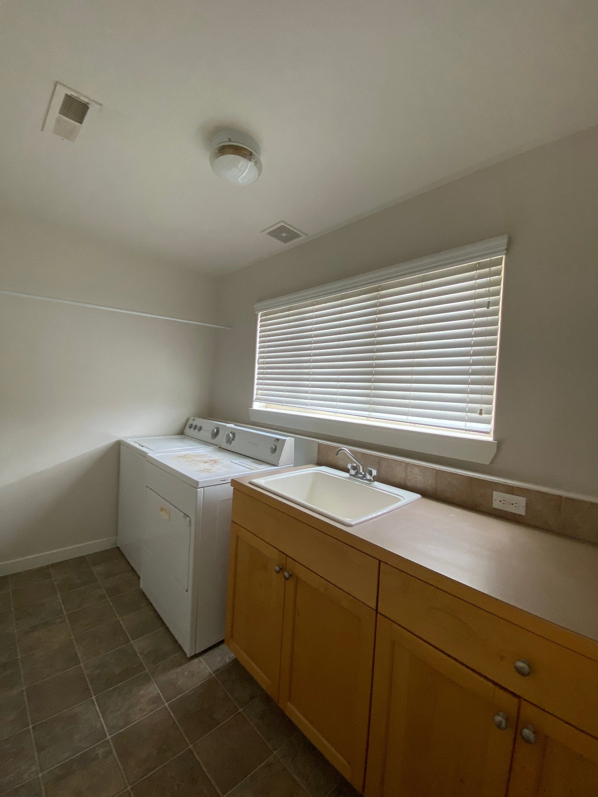 A laundry room with a white washer and dryer, a sink on a wooden cabinet, and a window with closed blinds.