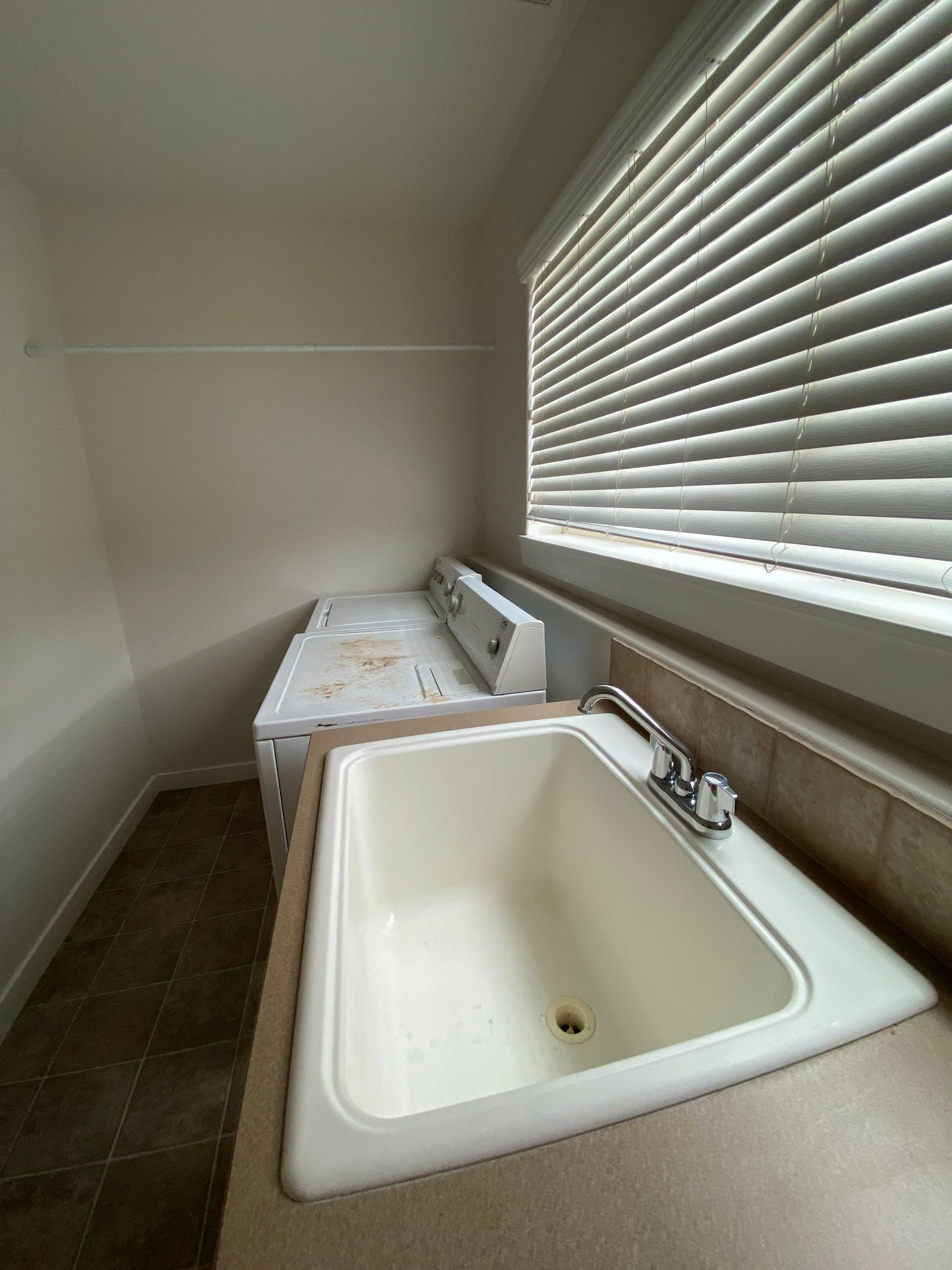 A utility sink sits beside a washer and dryer beneath a window with closed blinds.