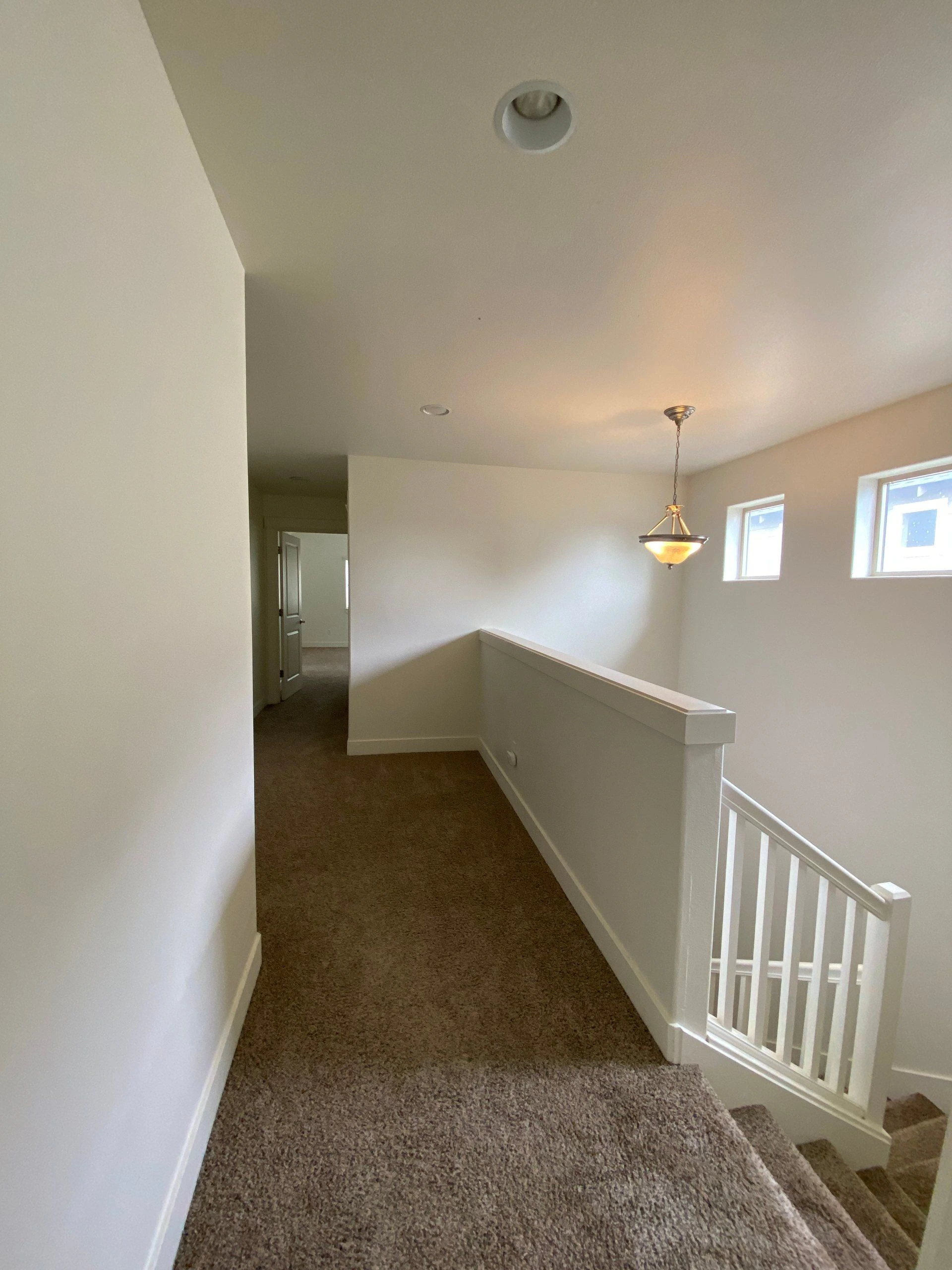 A carpeted upstairs landing has a white stair railing, two windows, a hanging light, and an open doorway.