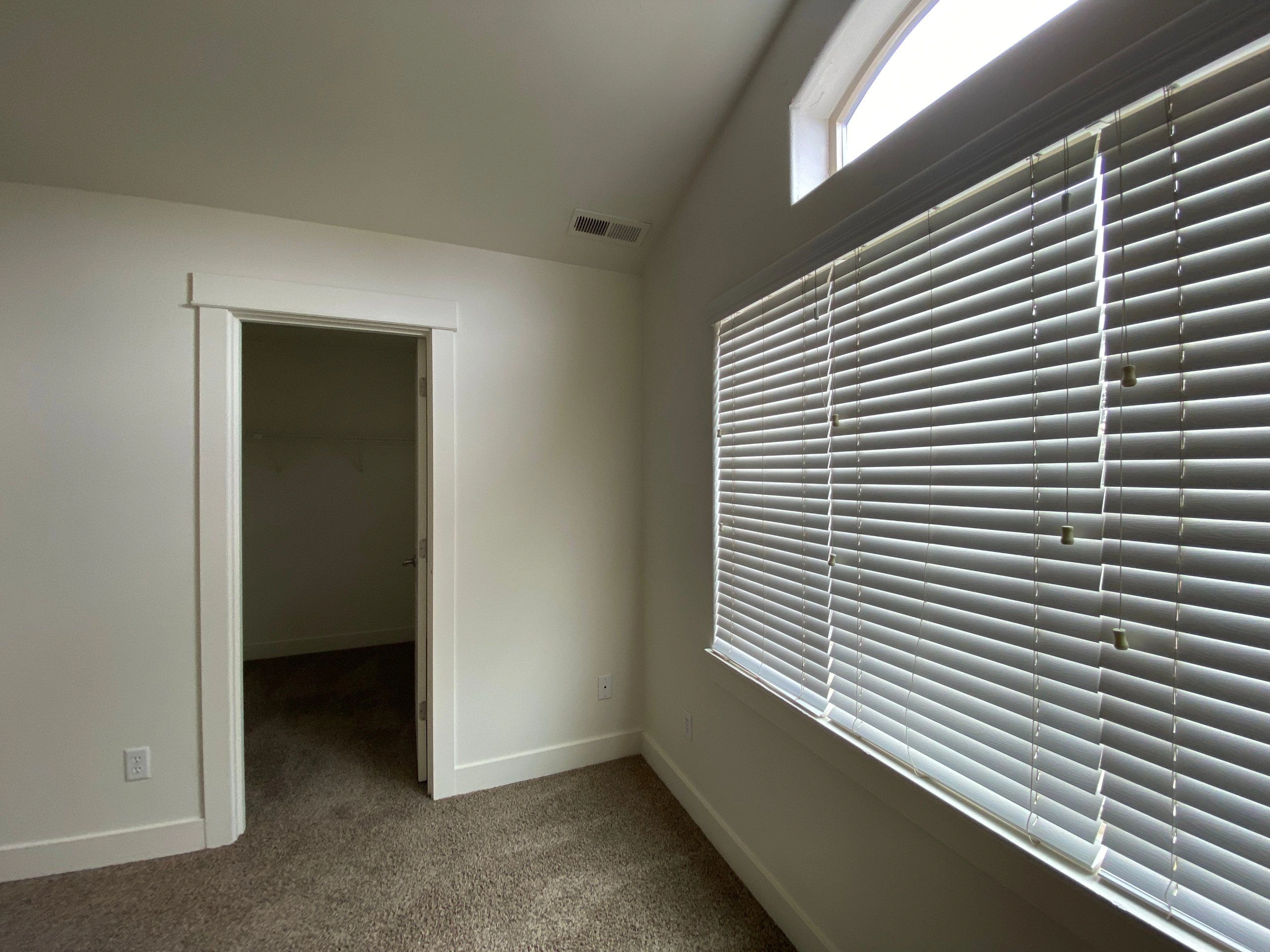 An empty carpeted room has white walls, an open doorway, and a large window covered by horizontal blinds.