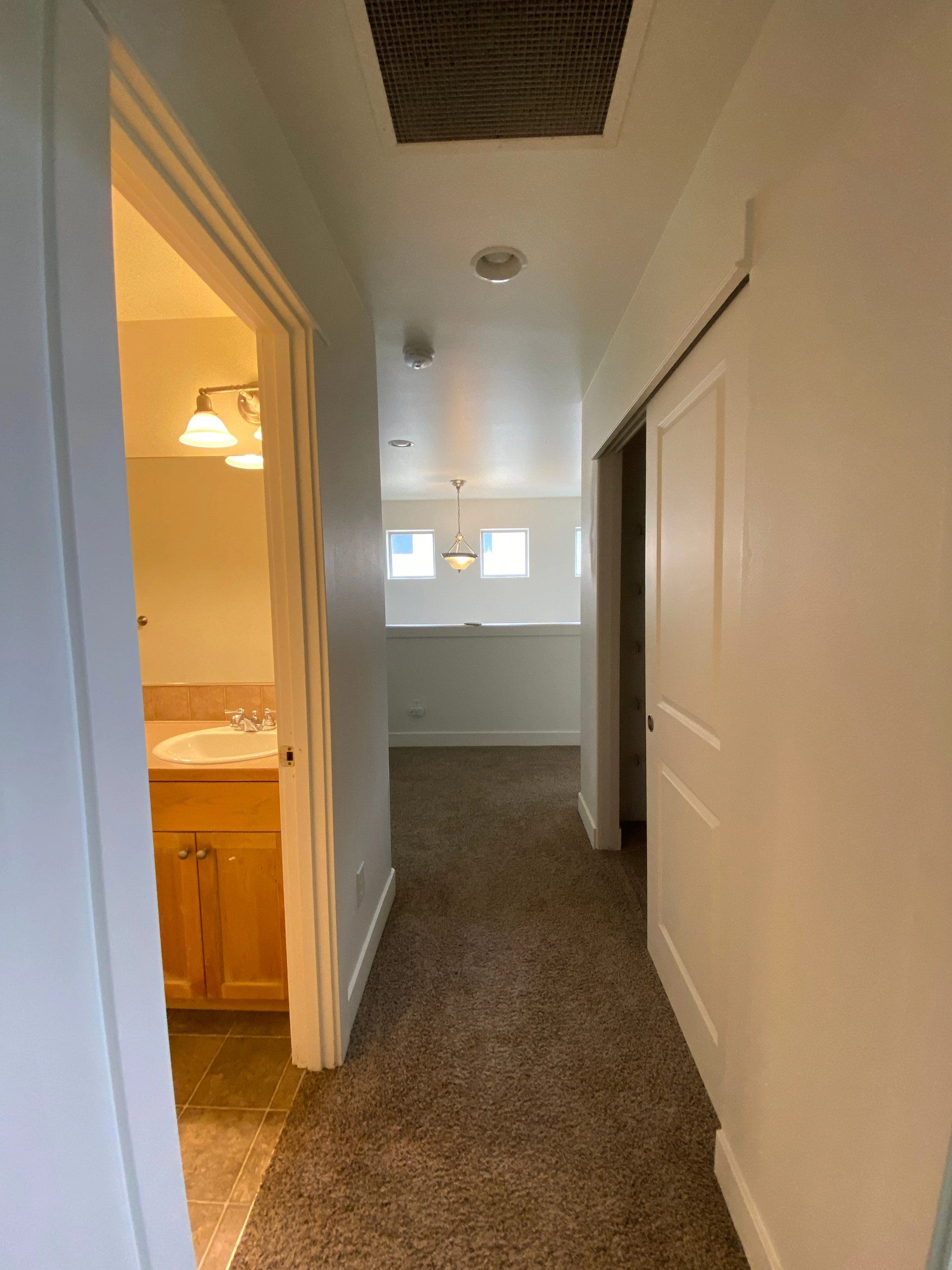 A carpeted hallway has an open bathroom doorway with a sink on the left, an open door on the right, and windows at the far end.