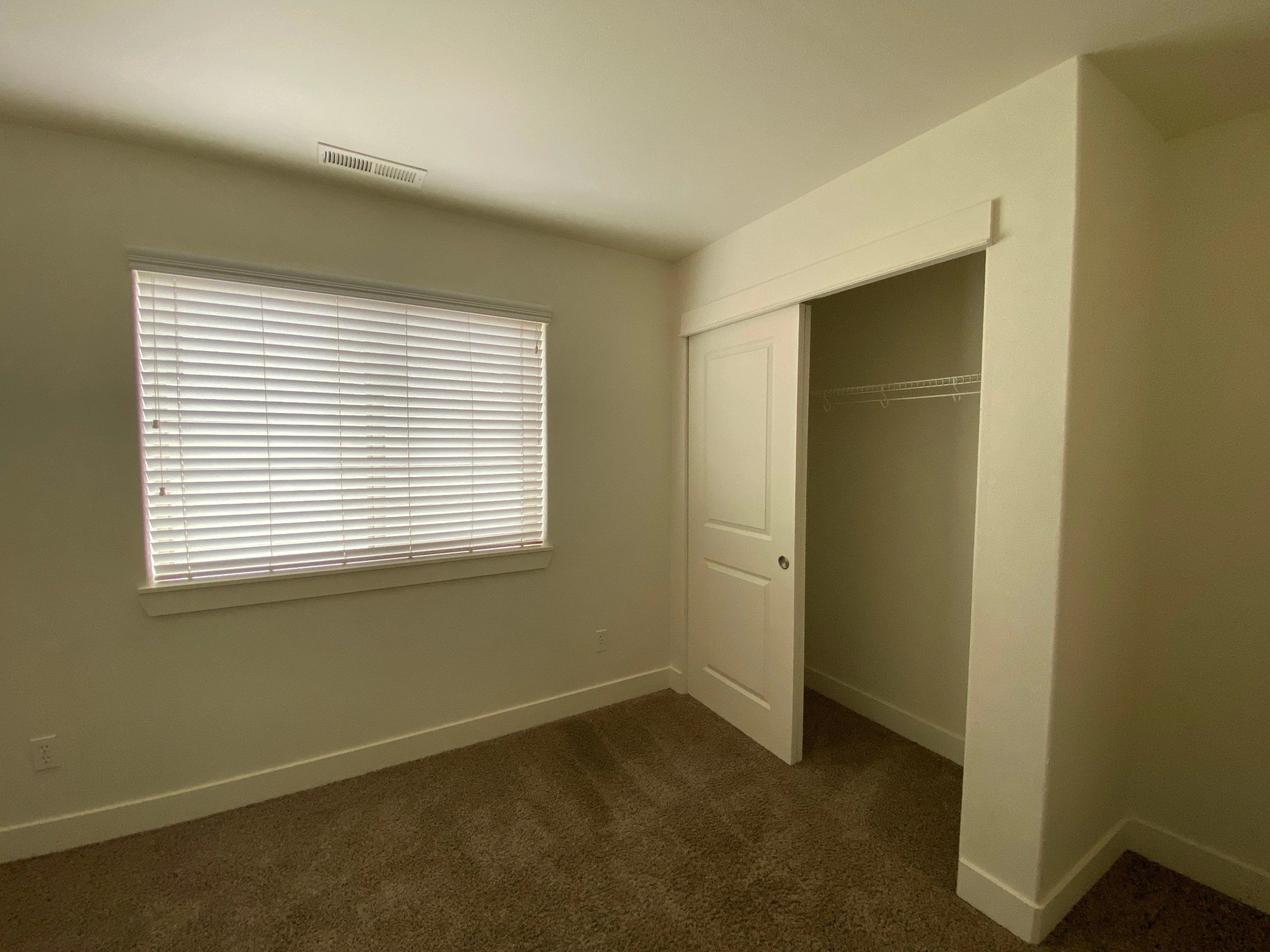 An empty carpeted room has a window with closed blinds and an open closet with a clothes rail.