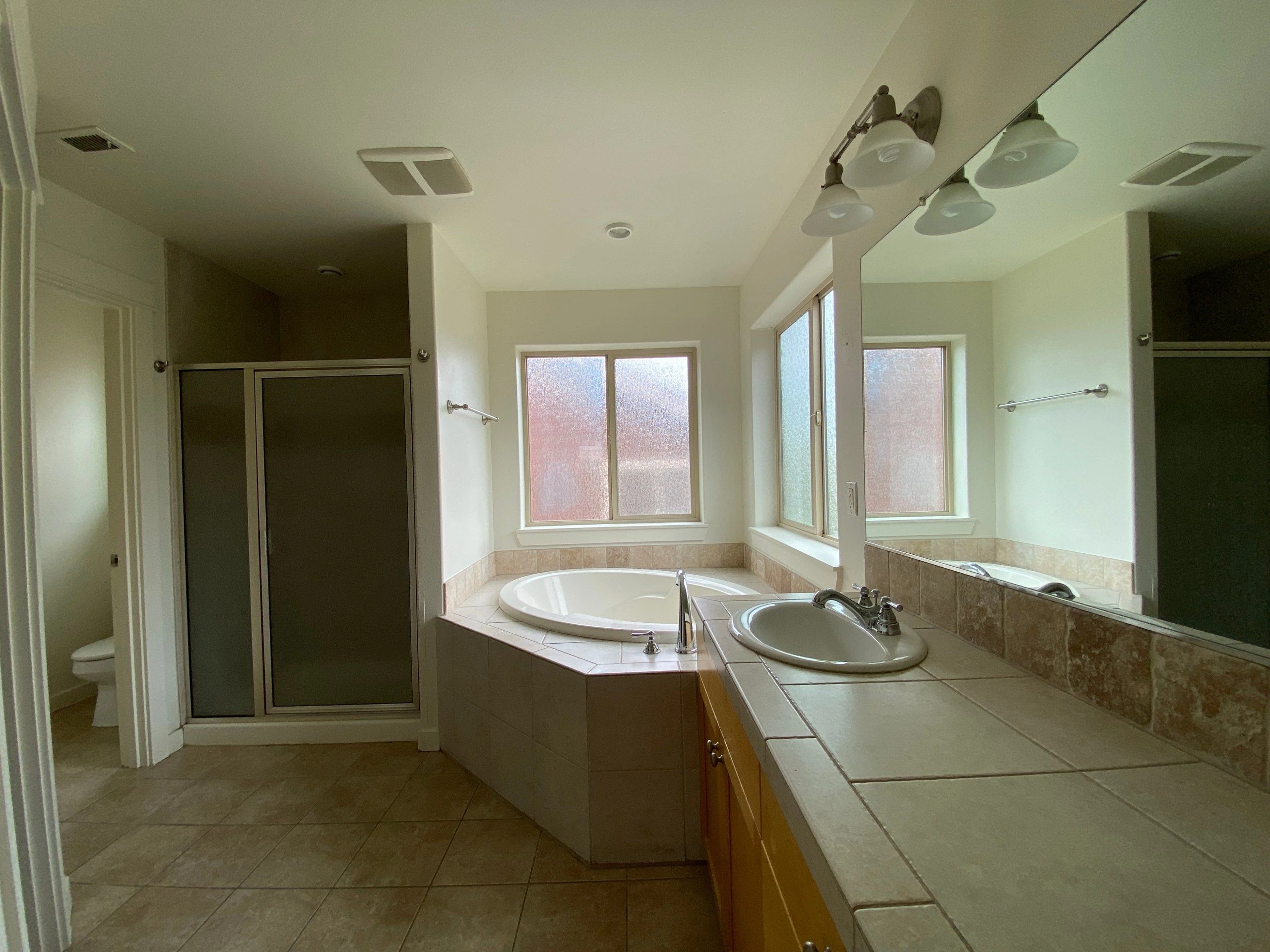 A bathroom with a corner bathtub, sink and large mirror, frosted windows, and a glass shower enclosure.