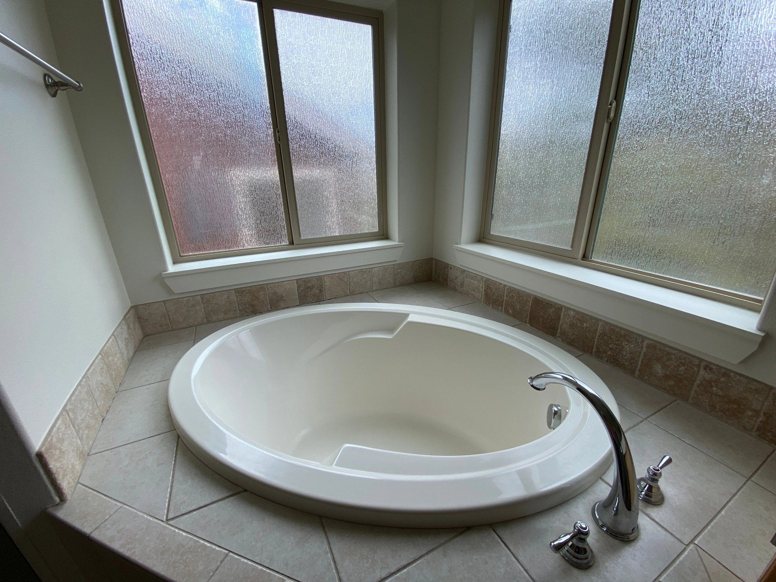A white oval bathtub sits beneath two sets of textured-glass windows, with beige tile around the tub and chrome fixtures.