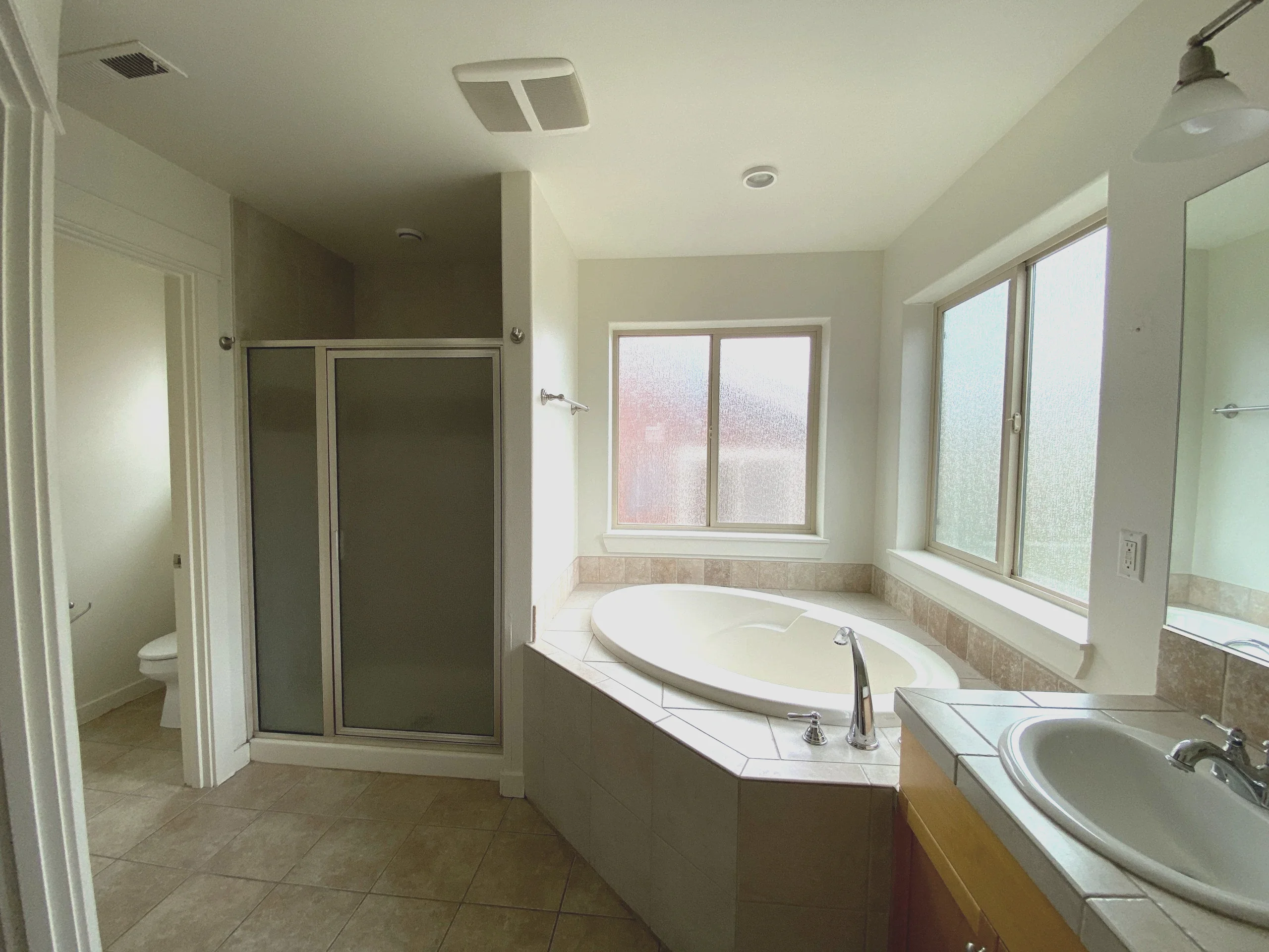 A tiled bathroom has a corner bathtub, frosted windows, a glass shower enclosure, and a sink; a toilet is visible through the doorway.