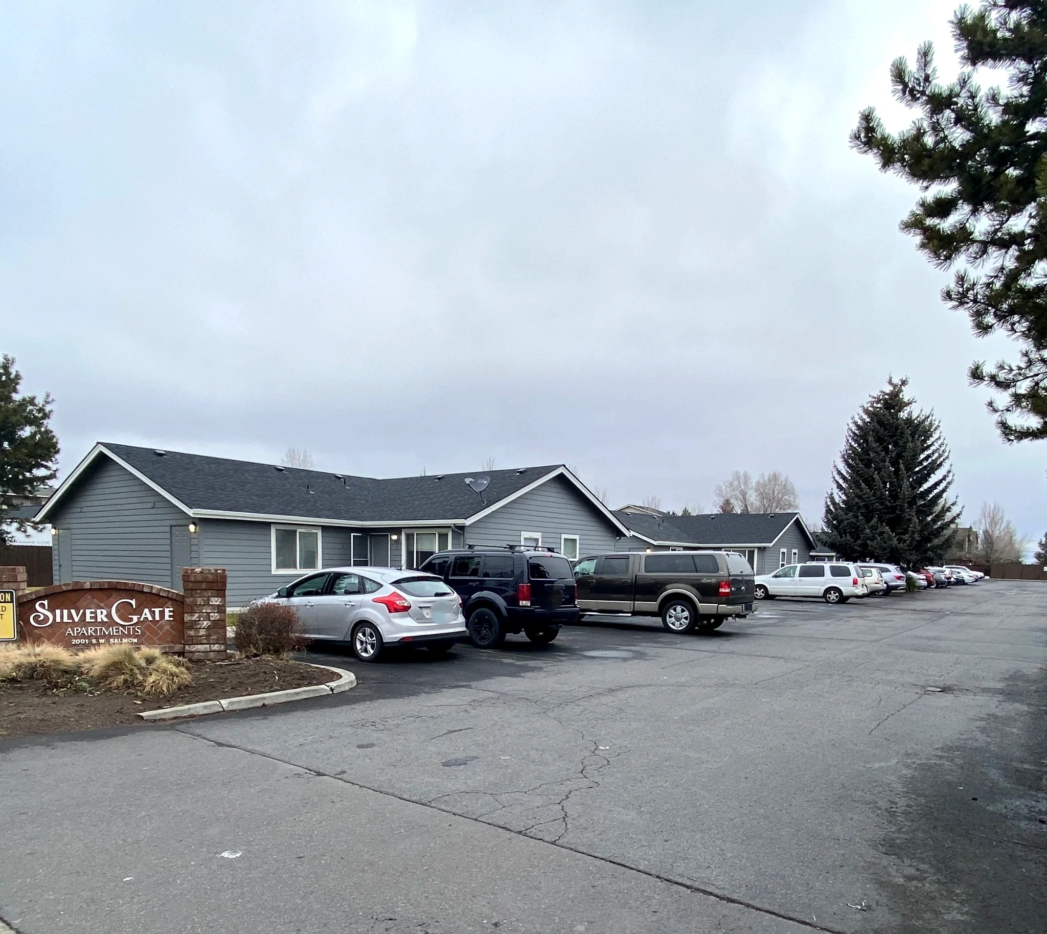 A gray, single-story building stands beside a parking lot with several cars and a sign reading “Silver Gate Apartments.”