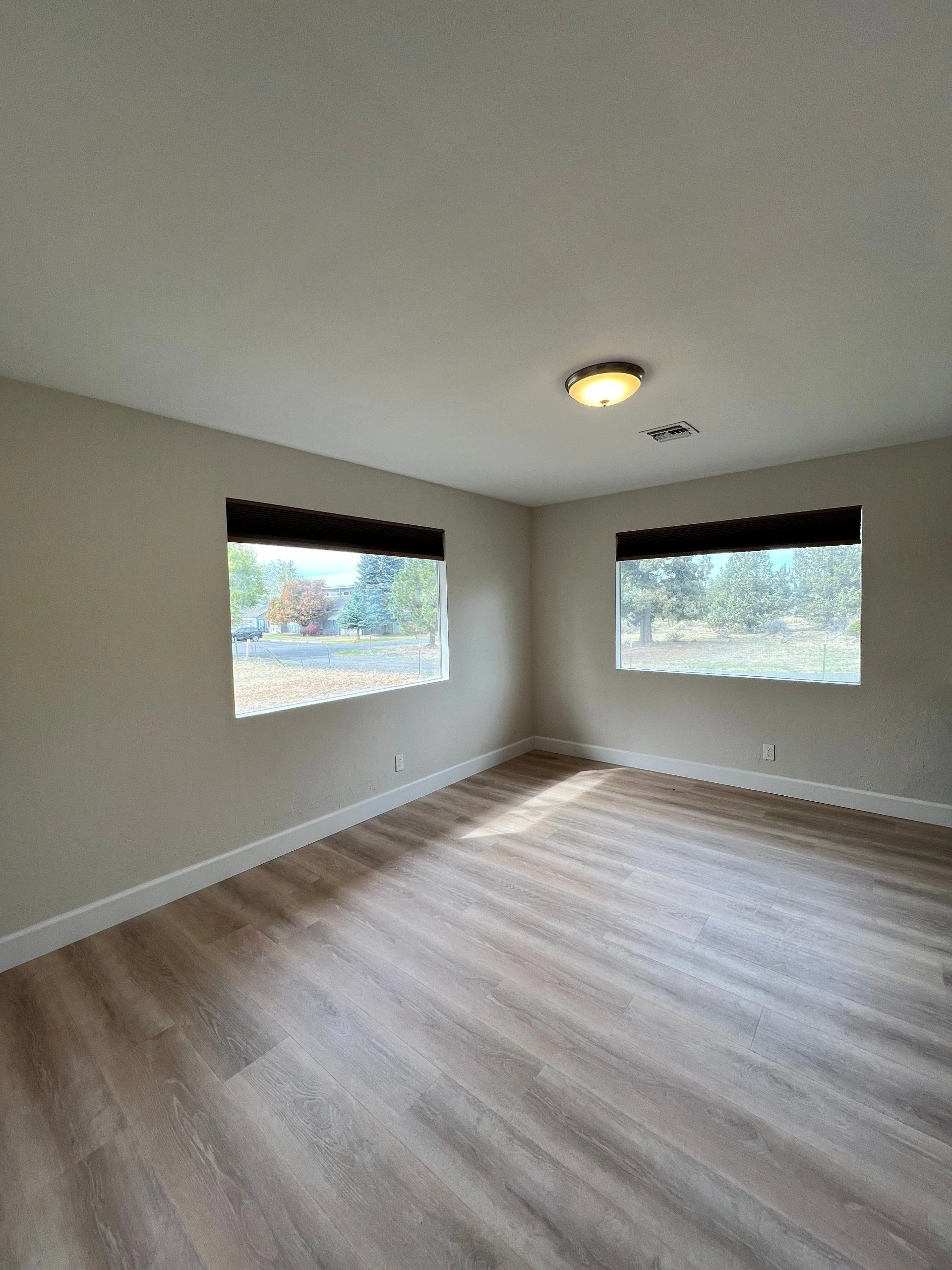 An empty room with two windows and dark blinds, gray-brown plank flooring, and a ceiling light and vent.