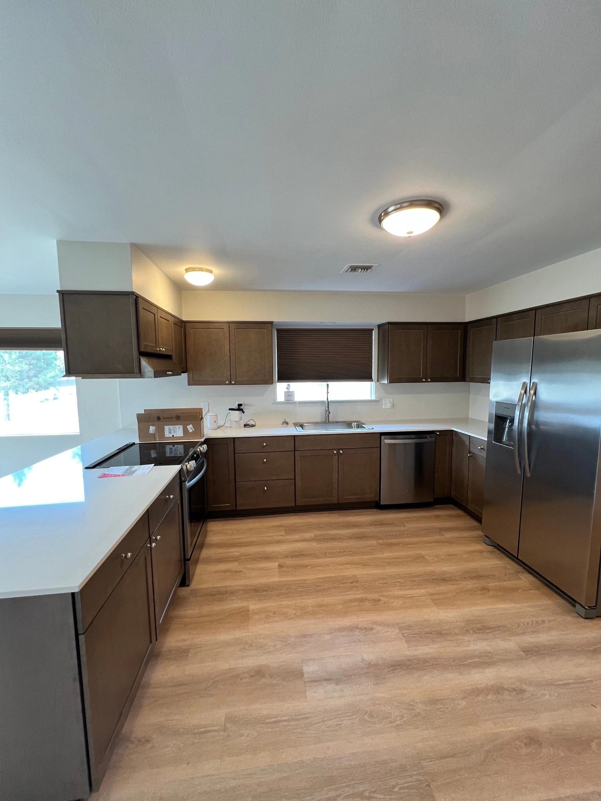 A kitchen with dark wood cabinets, white counters, a sink, stove, dishwasher, and stainless steel refrigerator.