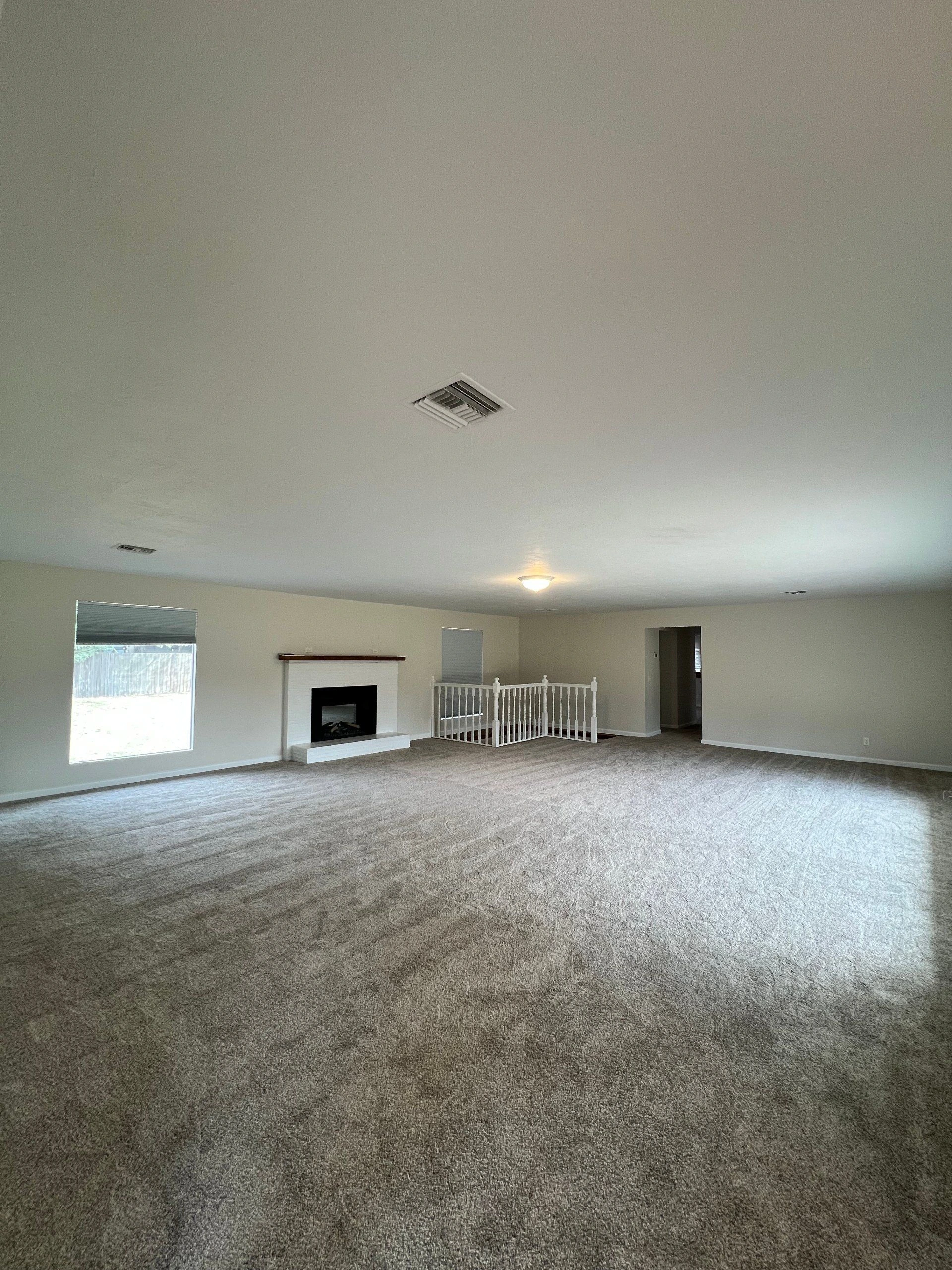 An empty carpeted room with a fireplace, two windows, a ceiling light, and a white railing around an opening.