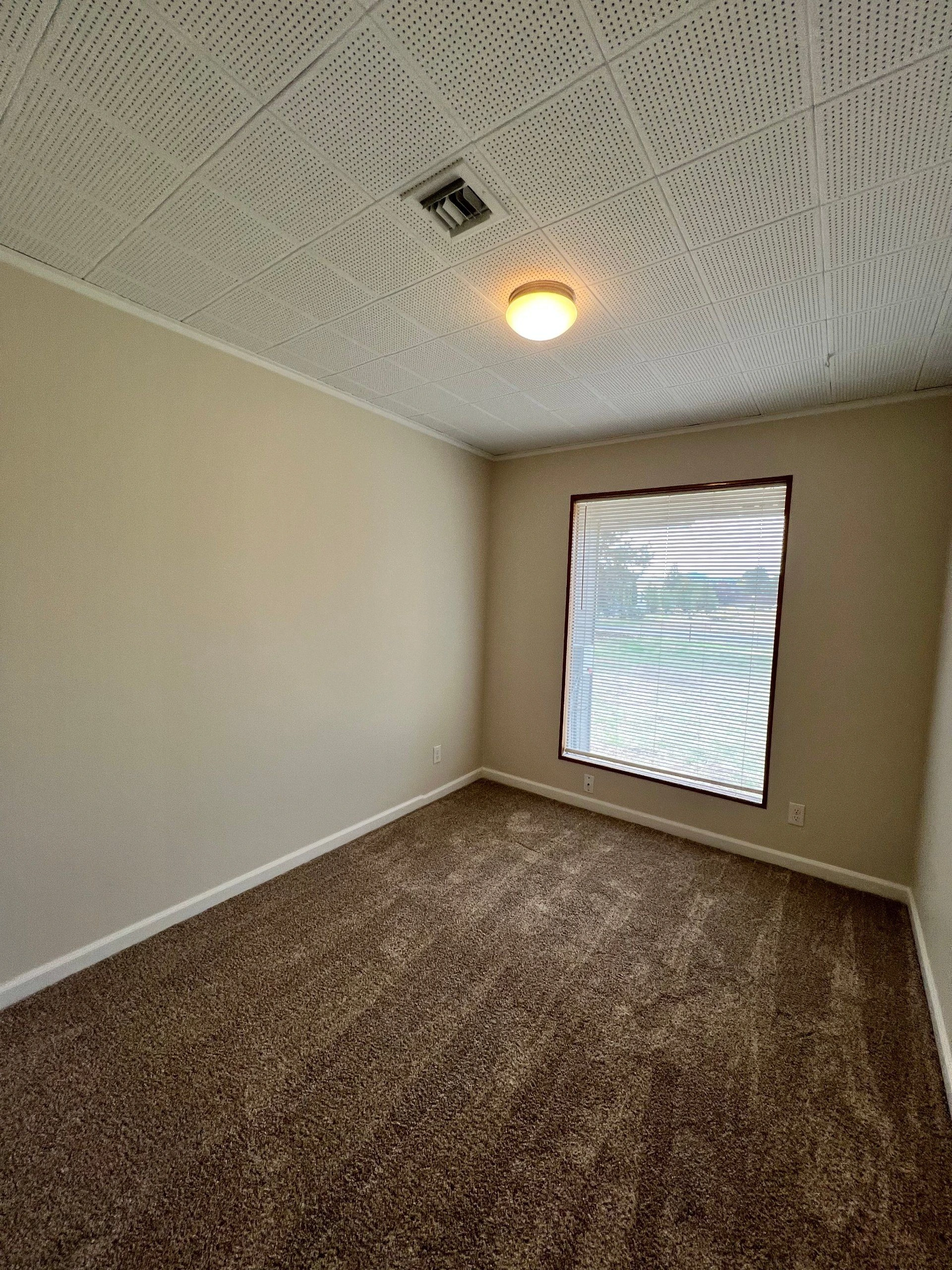 An empty room with brown carpet, beige walls, a lit ceiling fixture, and a window with closed blinds.