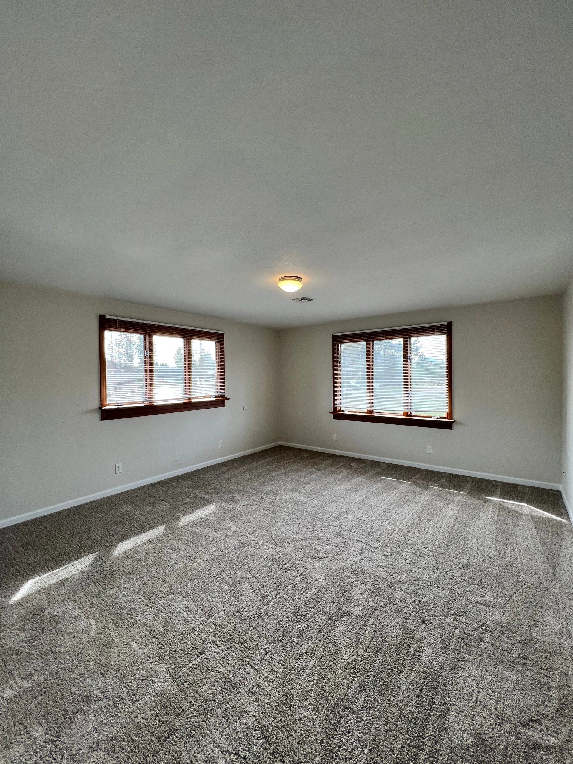 An empty room with gray carpet, two windows with wooden blinds, and a lit ceiling fixture.