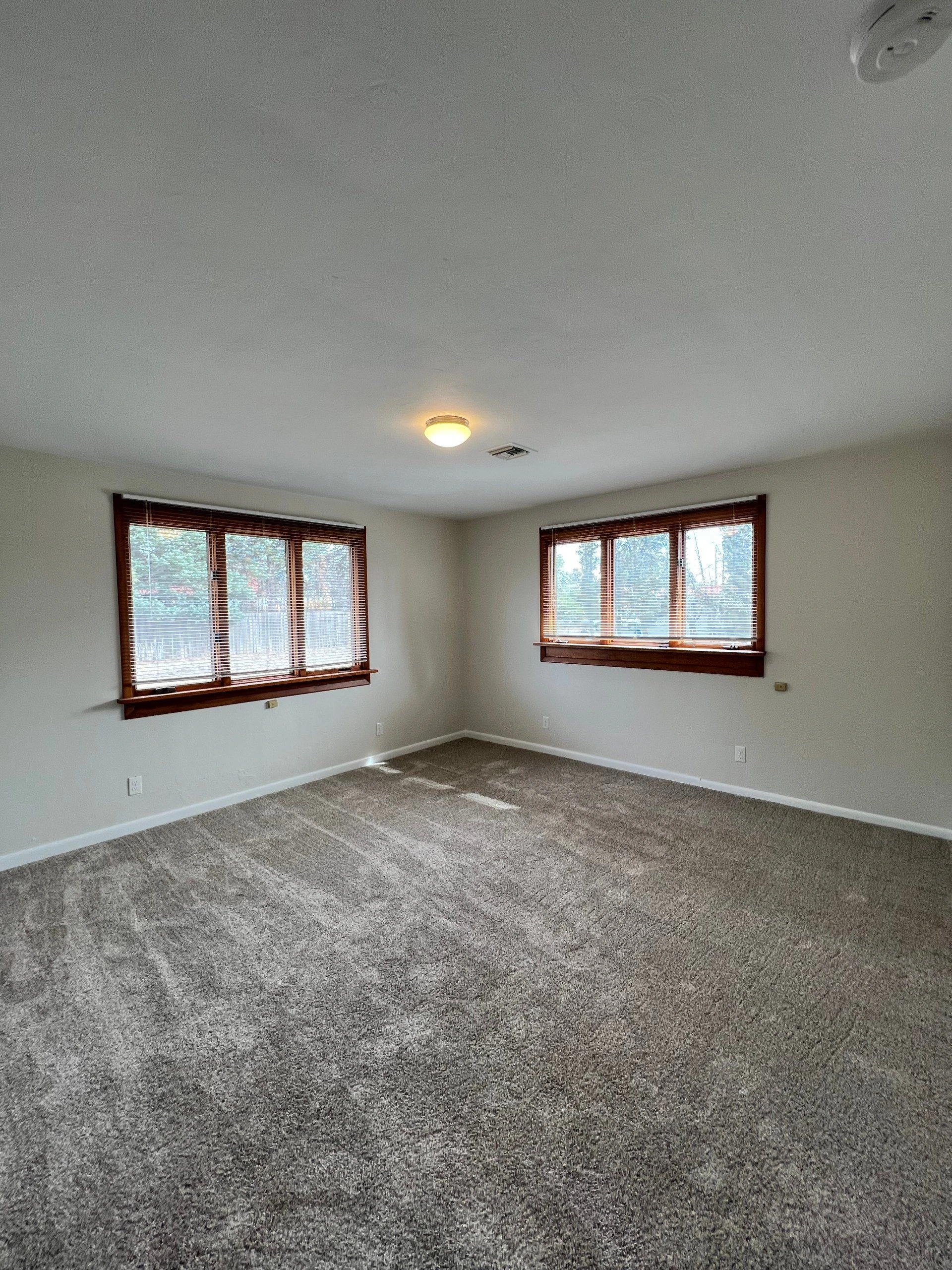 An empty room with gray carpet, pale walls, two windows with wooden blinds, and a lit ceiling fixture.