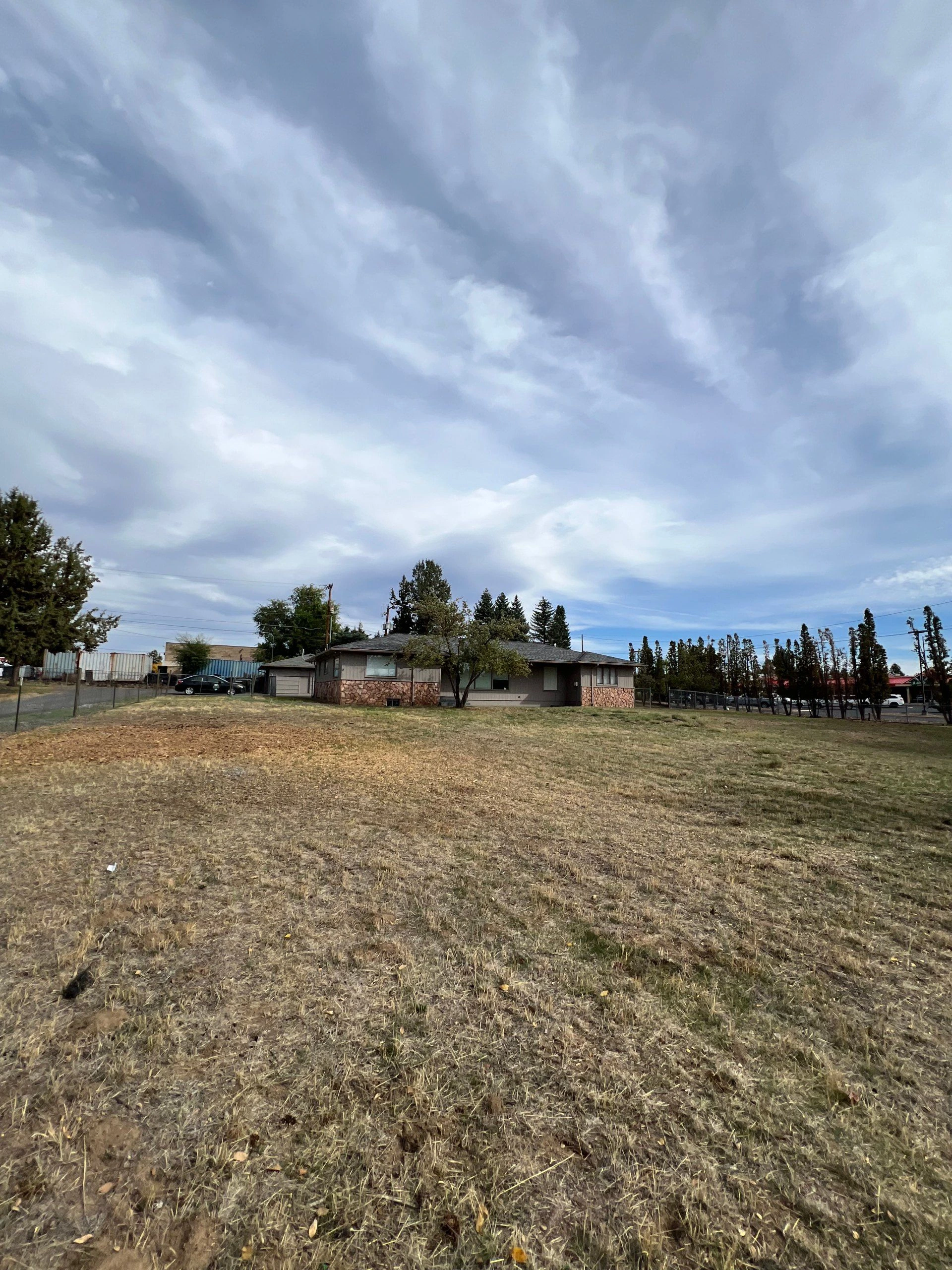 A single-story house stands beyond a dry, uneven field beneath a cloudy sky, with trees and parked vehicles nearby.