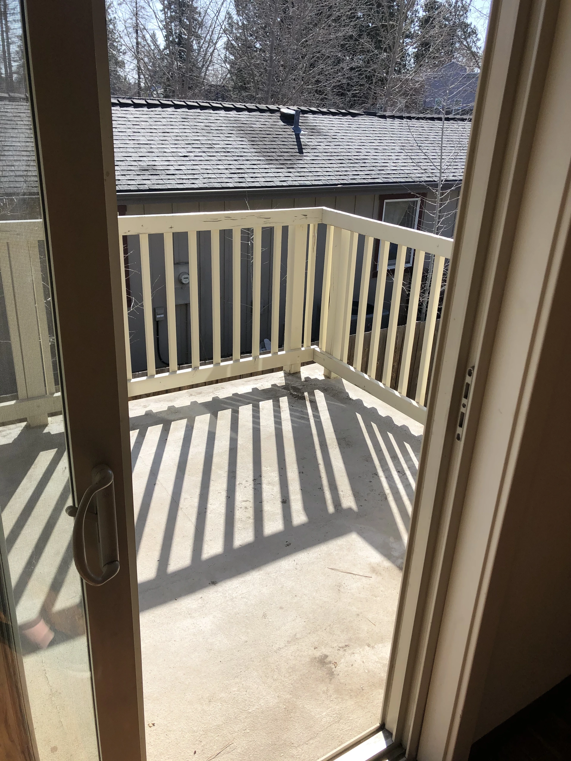 An open door leads to a concrete balcony with pale railings, casting long shadows; trees and a shingled roof are visible beyond.