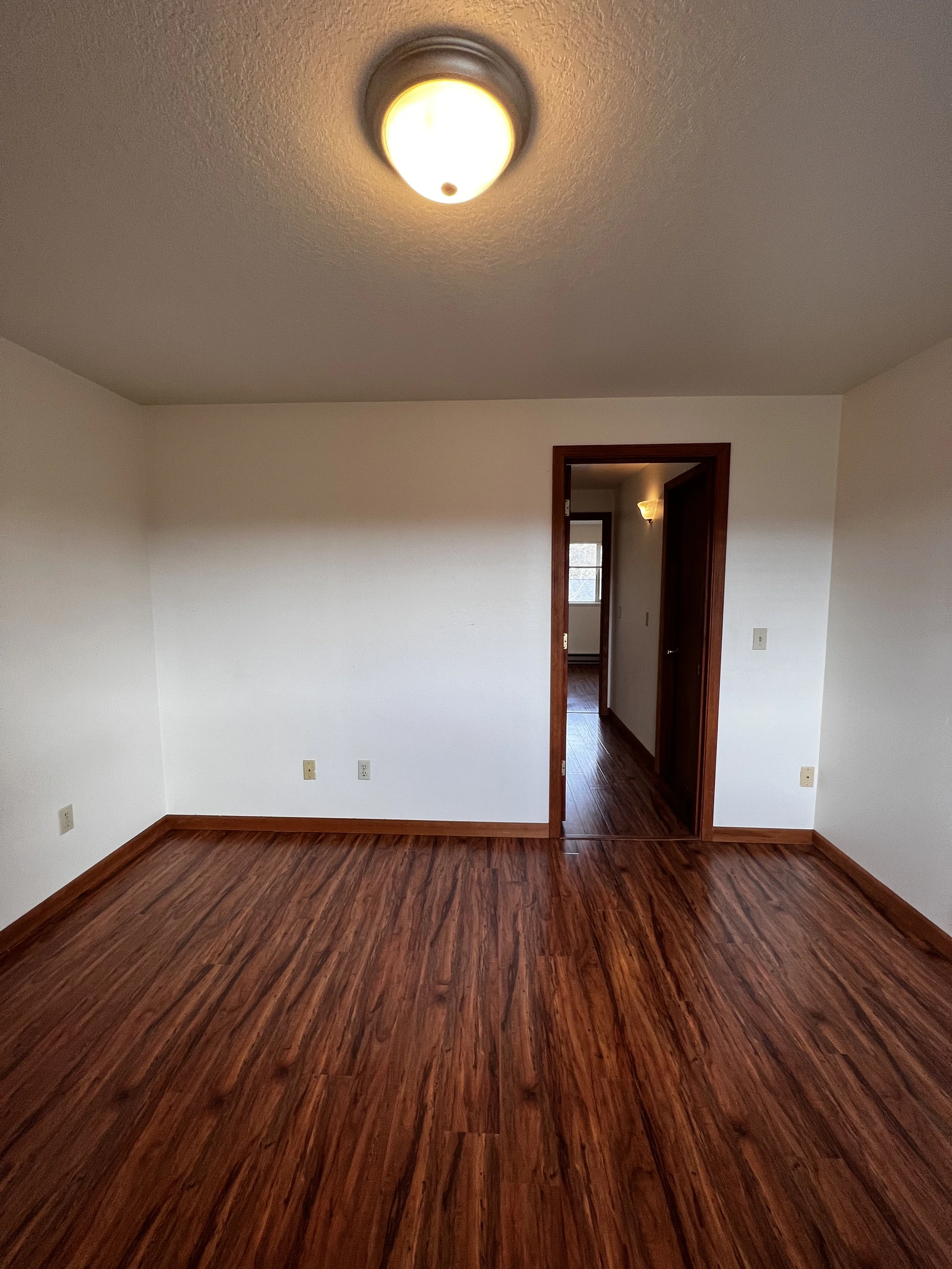 Empty room with dark wood flooring, white walls, a ceiling light, and an open doorway to a hallway.