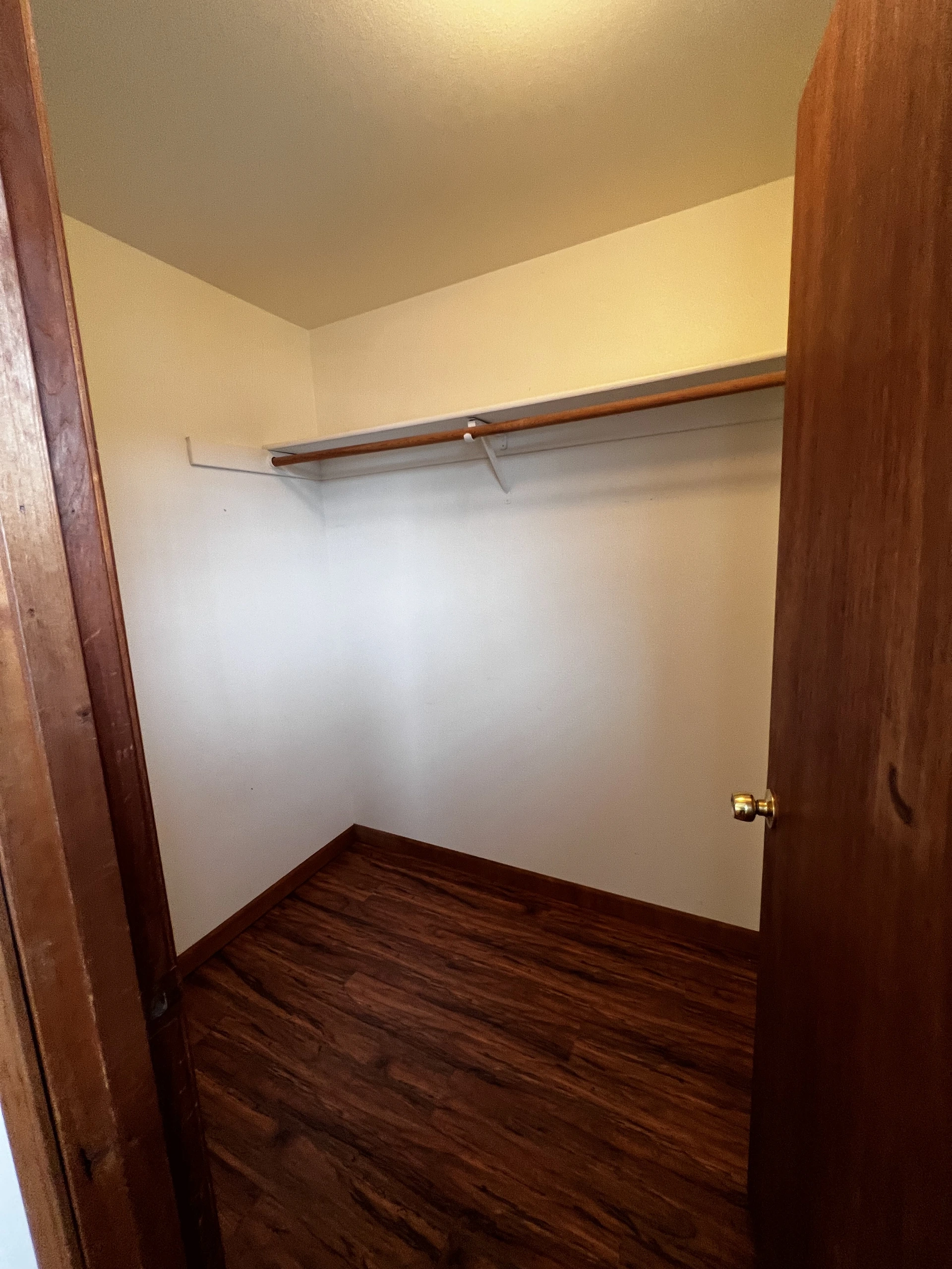 Open wooden door reveals an empty room with light walls, dark wood flooring, and a wall-mounted wooden rod beneath a shelf.
