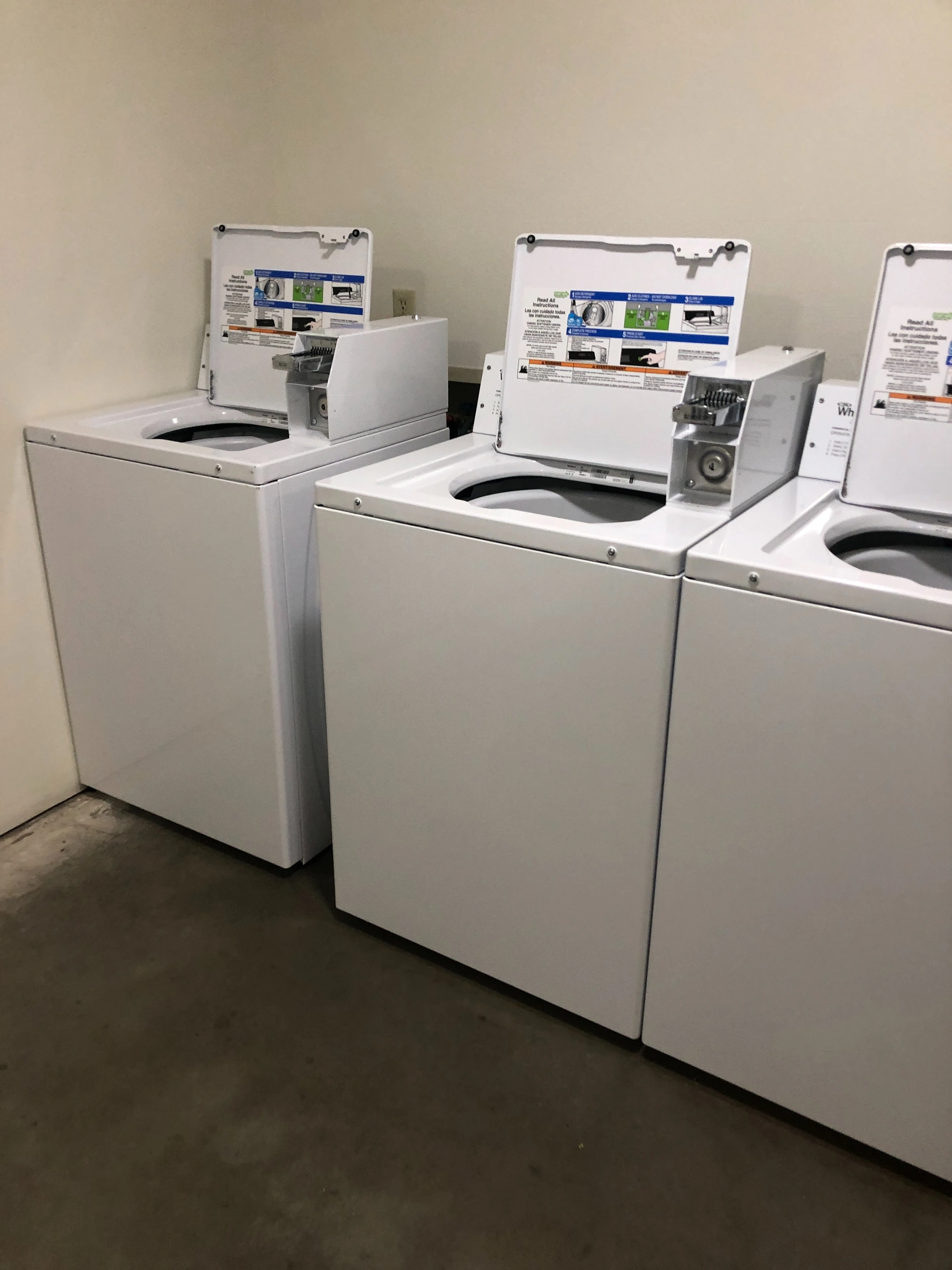 Three white top-loading washing machines with open lids and instruction panels stand against a beige wall.
