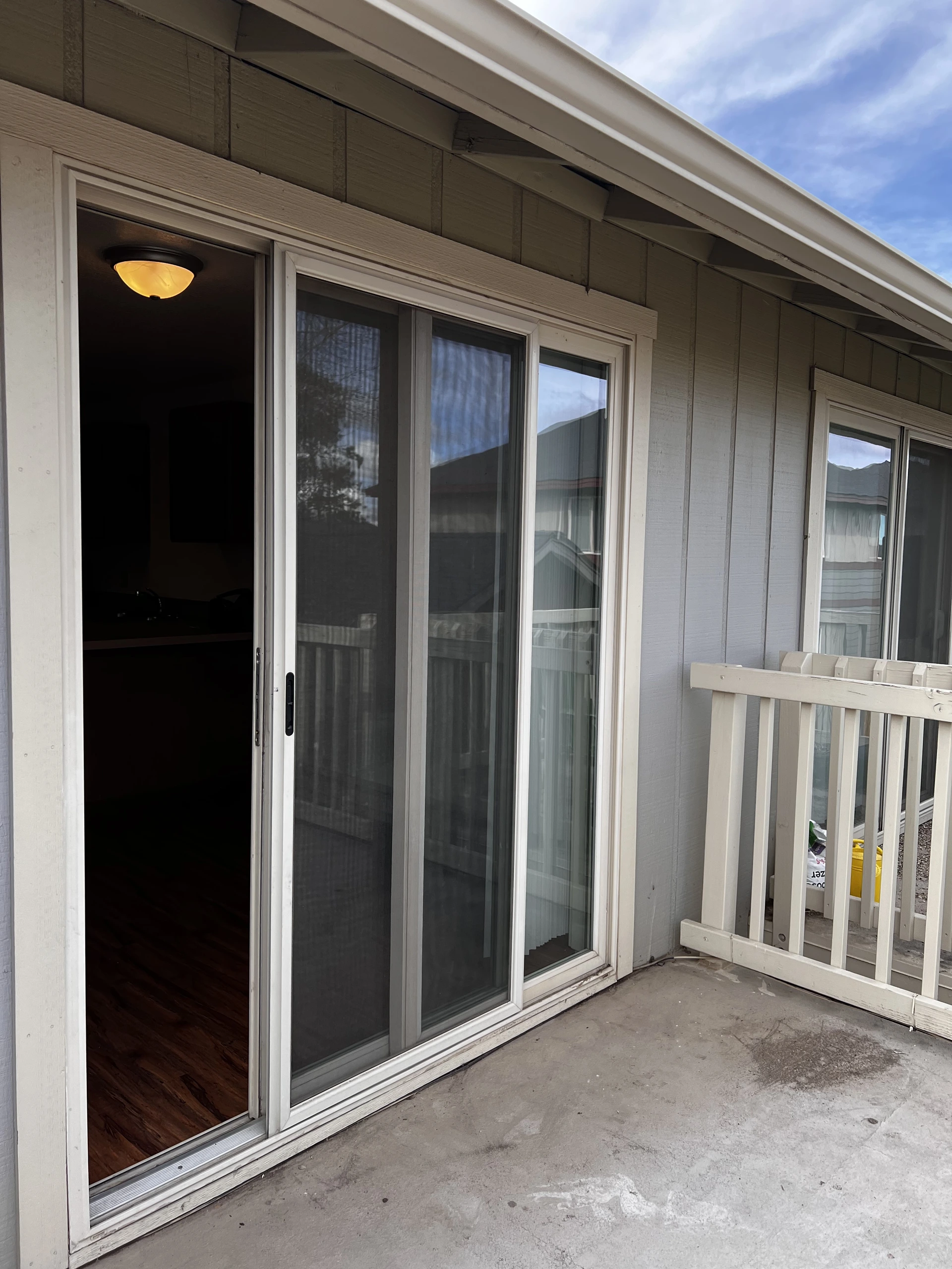 A concrete patio has an open sliding glass door, large windows, and a white railing beside gray siding.