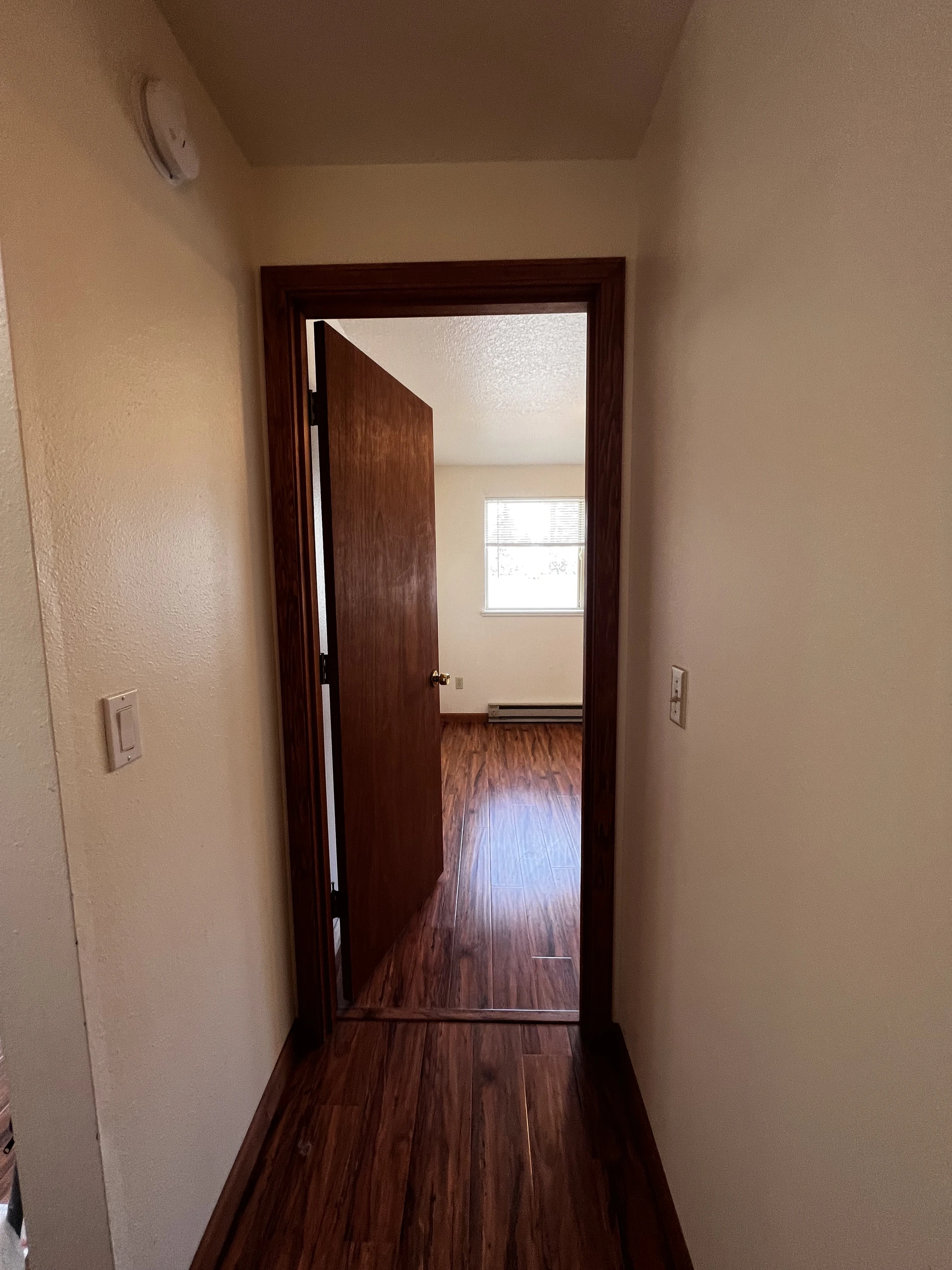 A narrow hallway with dark wood flooring leads to an open wooden door and a room with a window and blinds.
