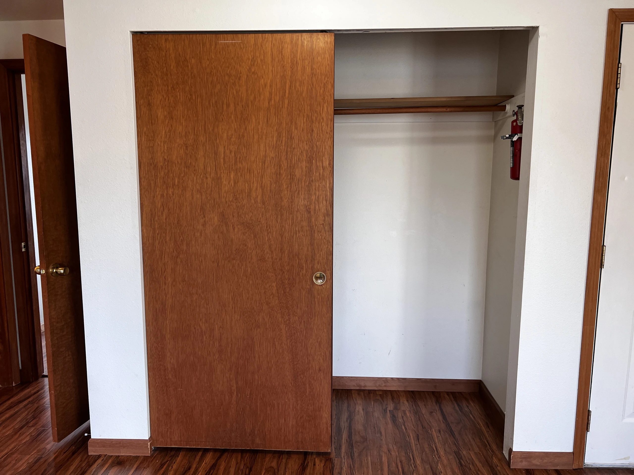 Open wooden closet with shelves, a hanging rod, and a red fire extinguisher; a sliding wooden door is partly closed.